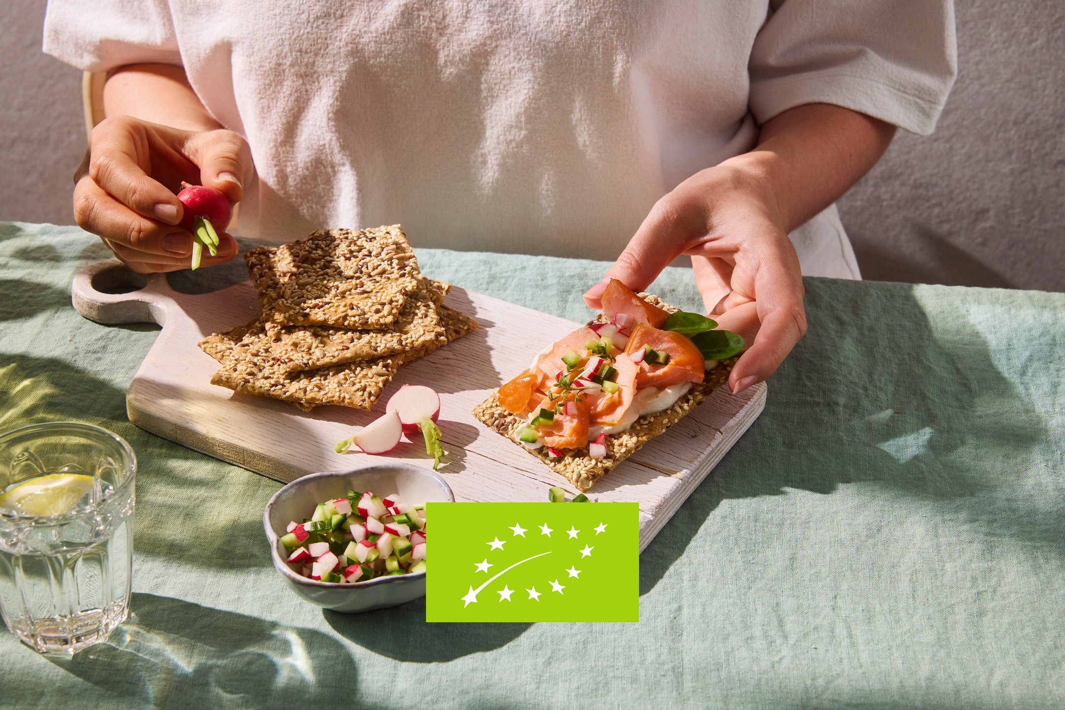 Hands preparing crispbread with salmon, cream cheese, and vegetables on a wooden board, with an organic logo.