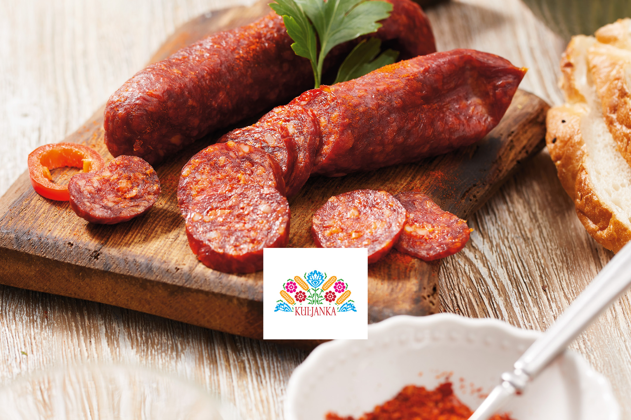 Sliced chorizo sausage and red pepper on a wooden board with a folk-art logo.