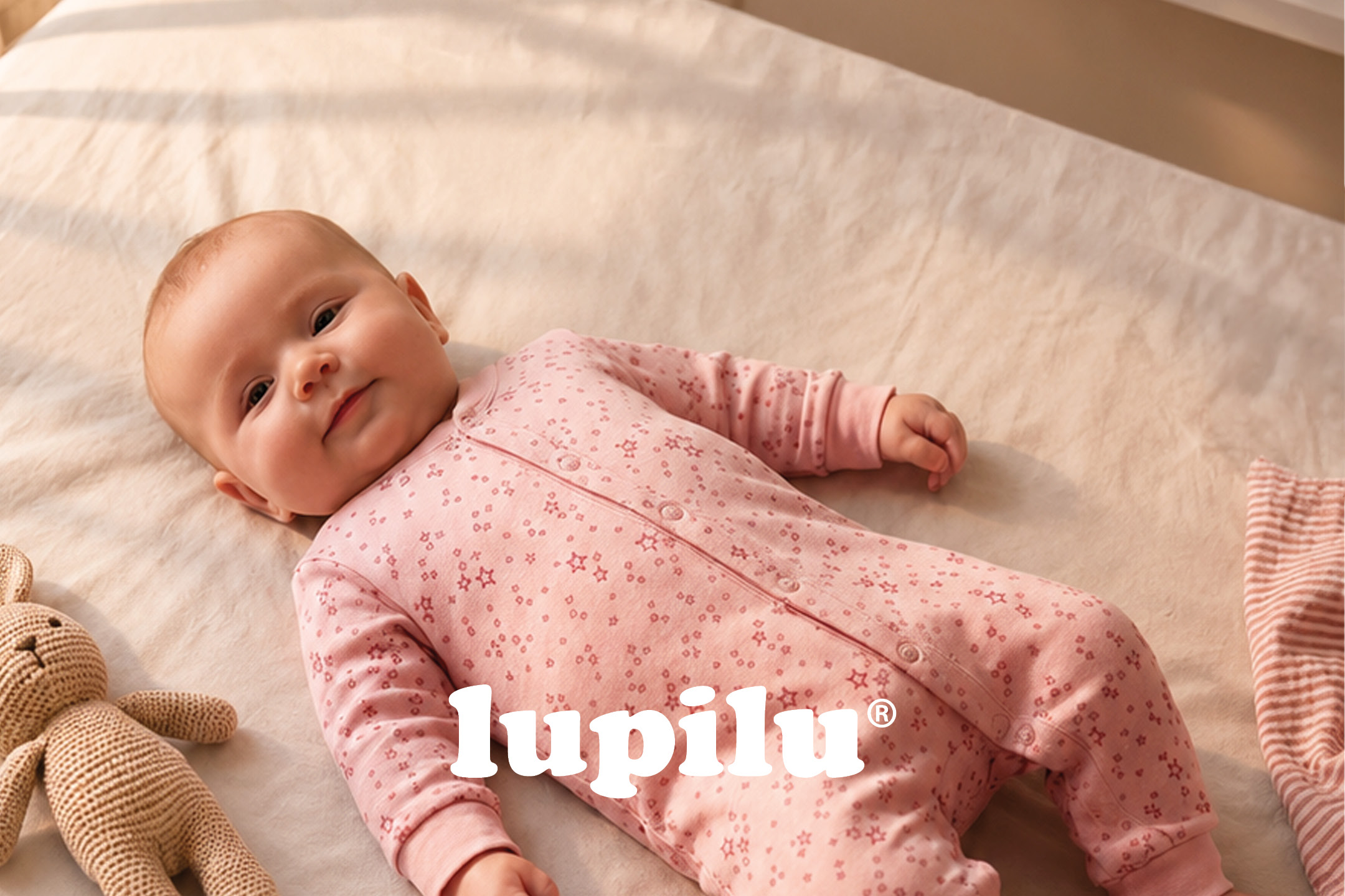 A baby in a pink star-patterned sleepsuit lies on a light-colored blanket.