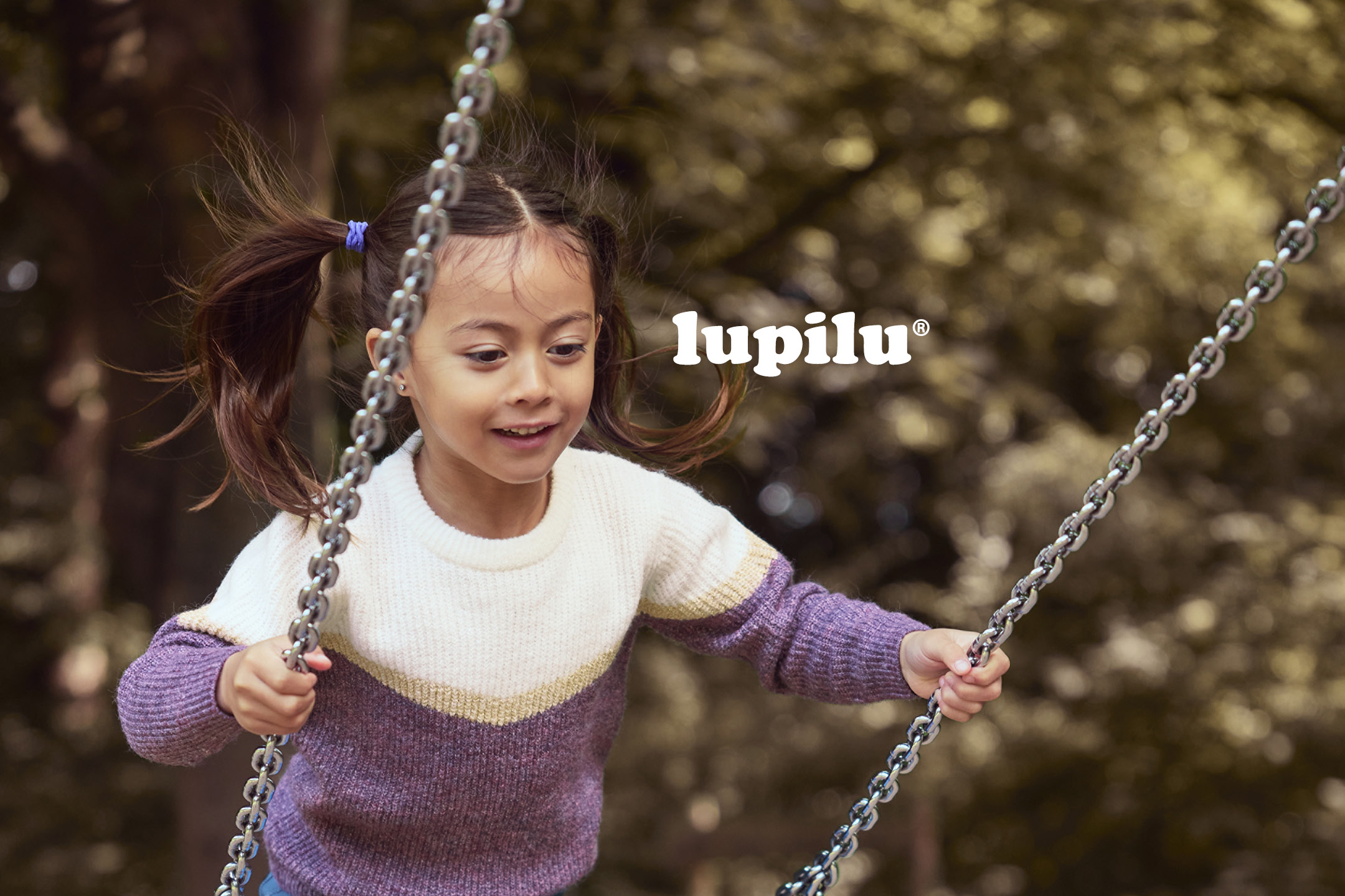 A smiling young girl in a white and purple knit sweater playing on a swing.