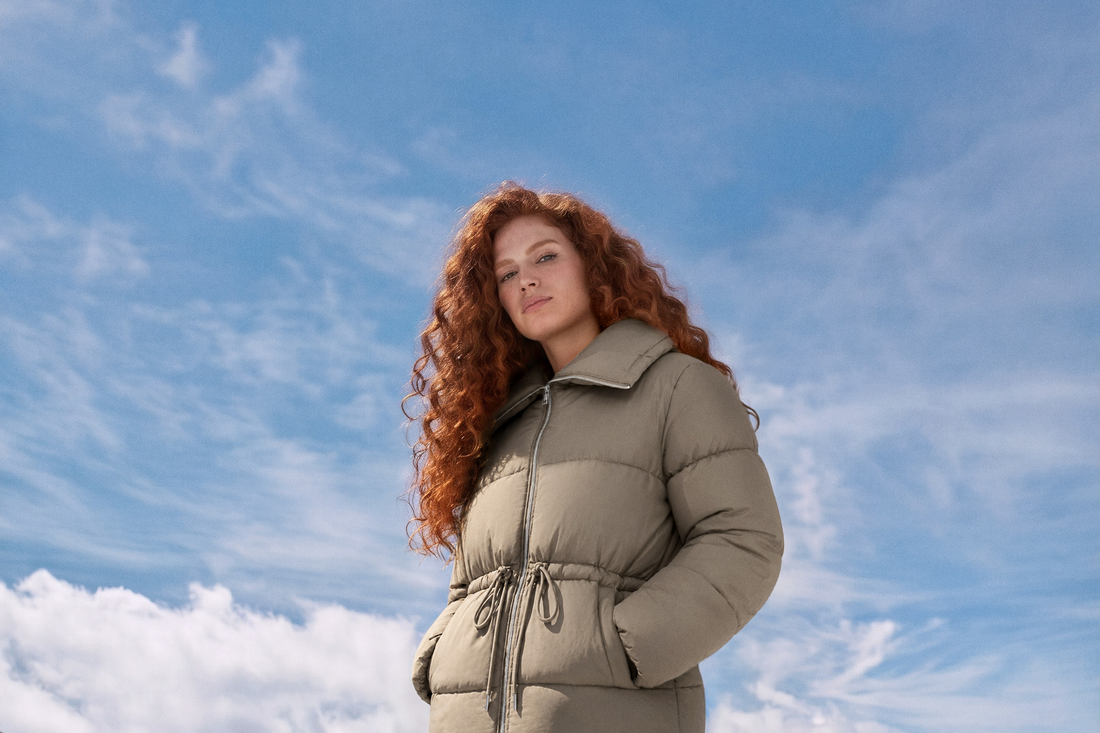 A woman with long curly red hair wearing a beige puffer jacket against a blue sky.