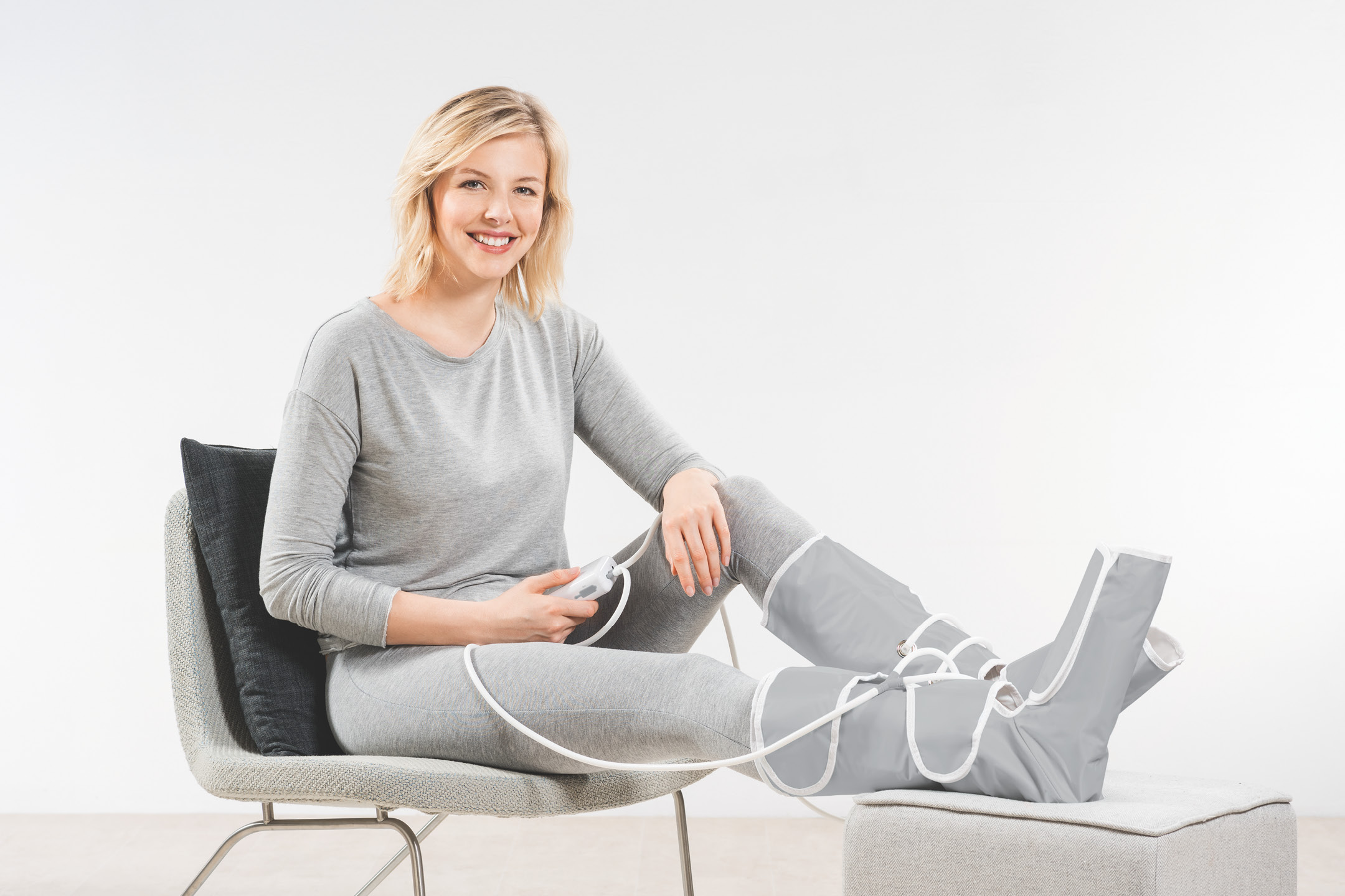 A person sitting in a chair using a grey leg compression massager with a remote control.