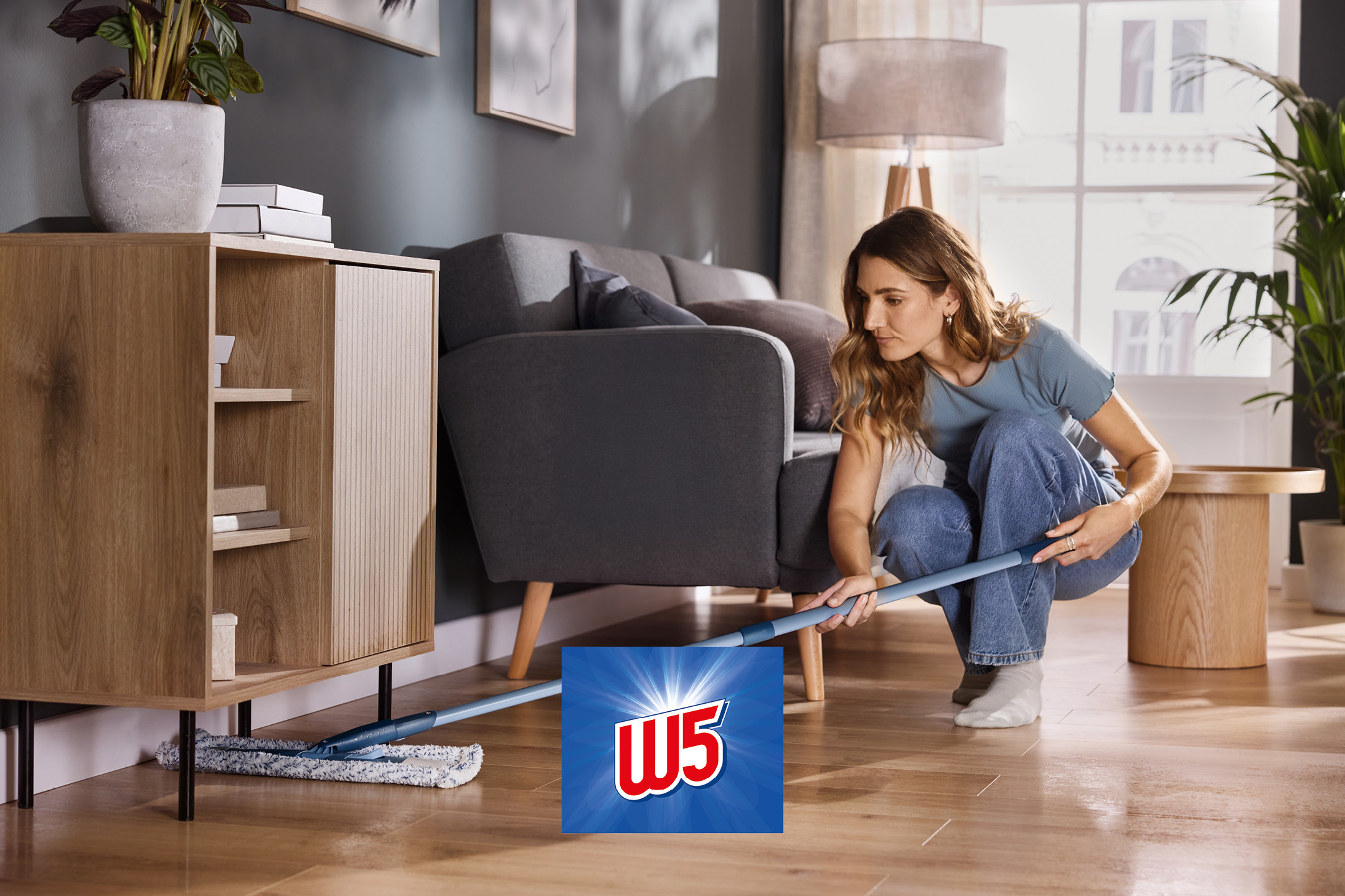 A person mopping a wooden floor under a cabinet with a flat mop.