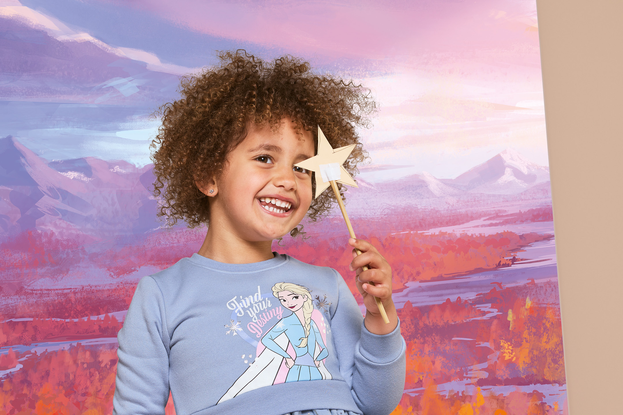 A smiling child with curly hair holding a star-shaped wand against a painted background.