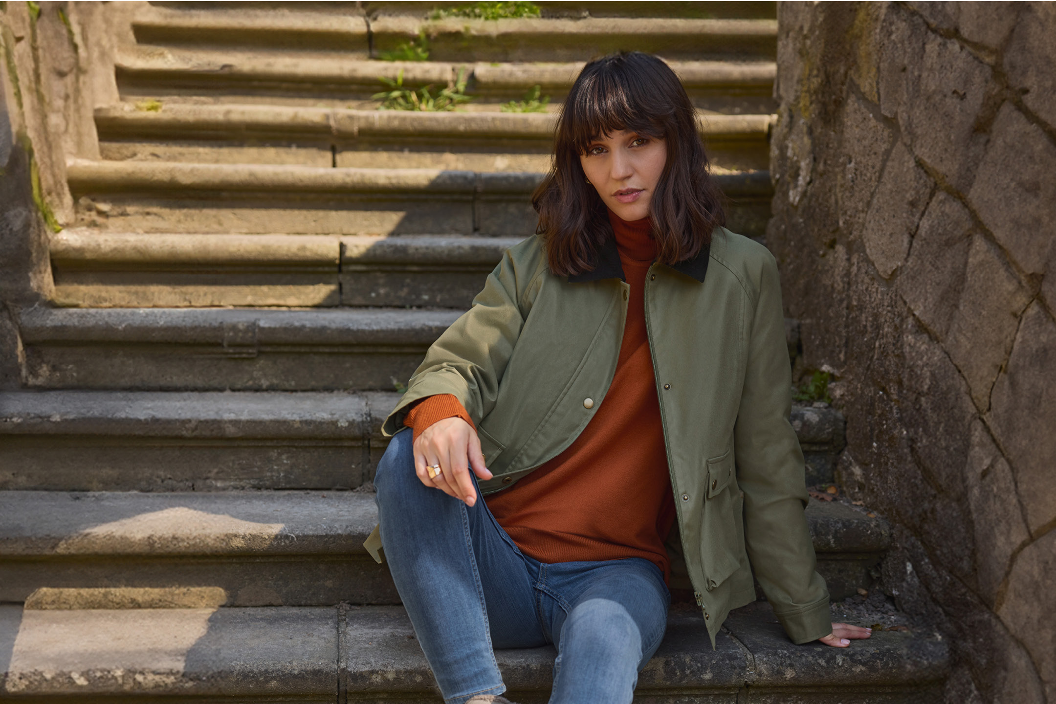 Woman in an olive green jacket, rust-colored turtleneck, and blue jeans sitting on stone steps.