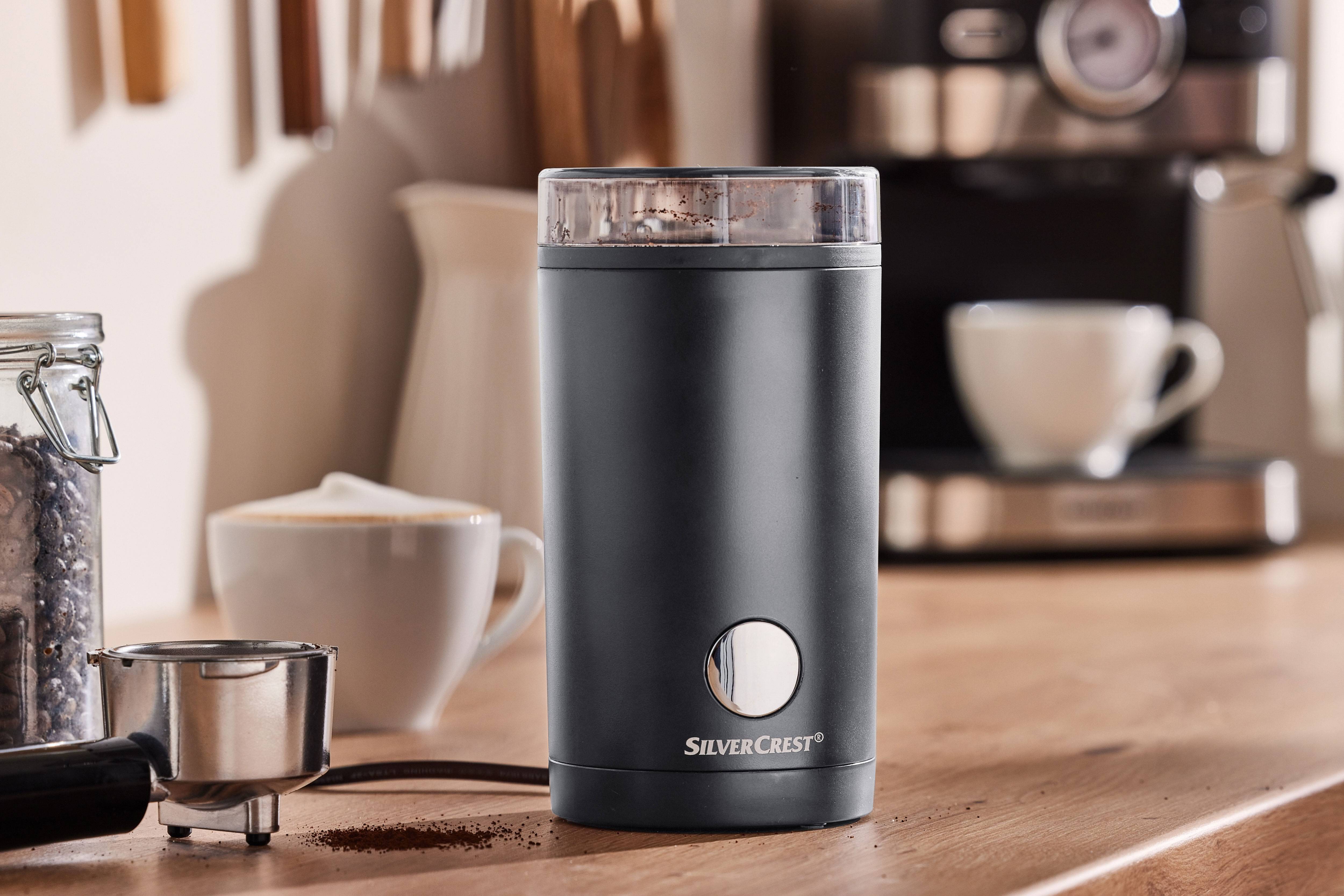 Black coffee grinder with ground coffee, a coffee machine, and cups on a wooden table.