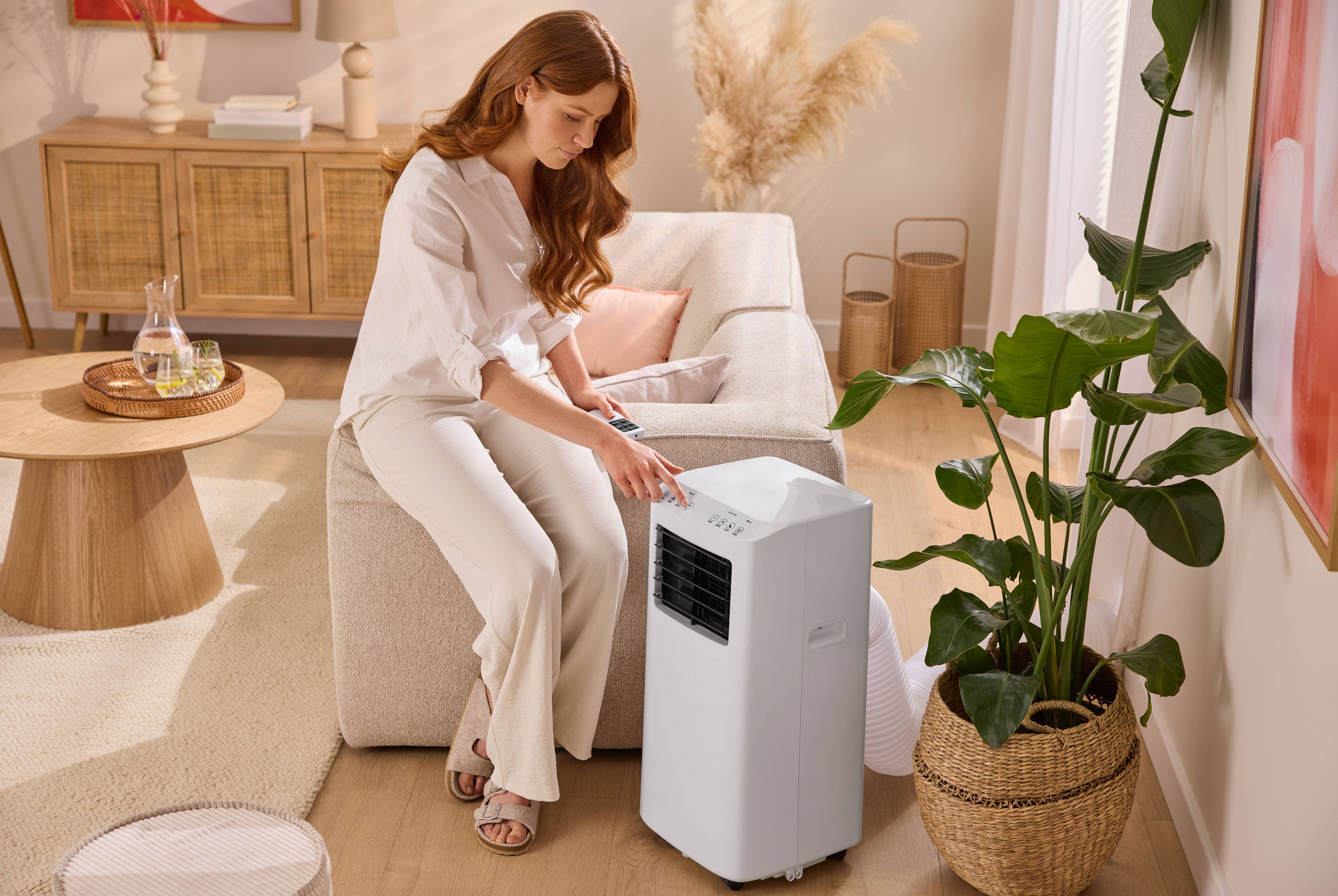 Woman sitting on a sofa, operating a portable air conditioner in a living room.
