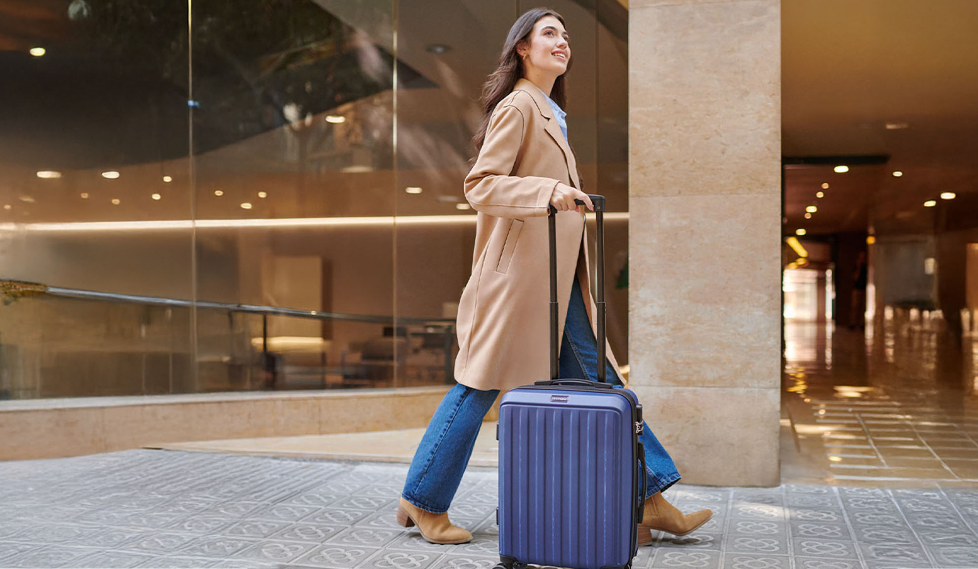 Woman in a beige coat and jeans pulling a blue suitcase on patterned pavement.