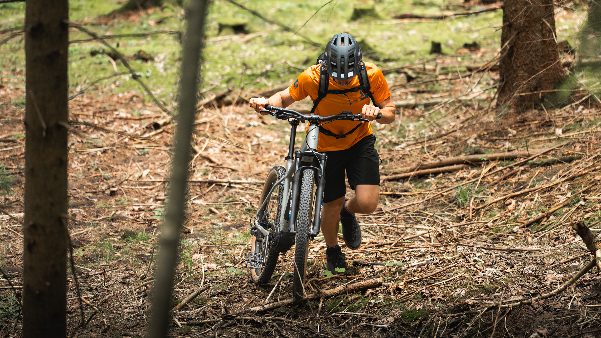 A cyclist in an orange shirt pushes an electric mountain bike through a forest.