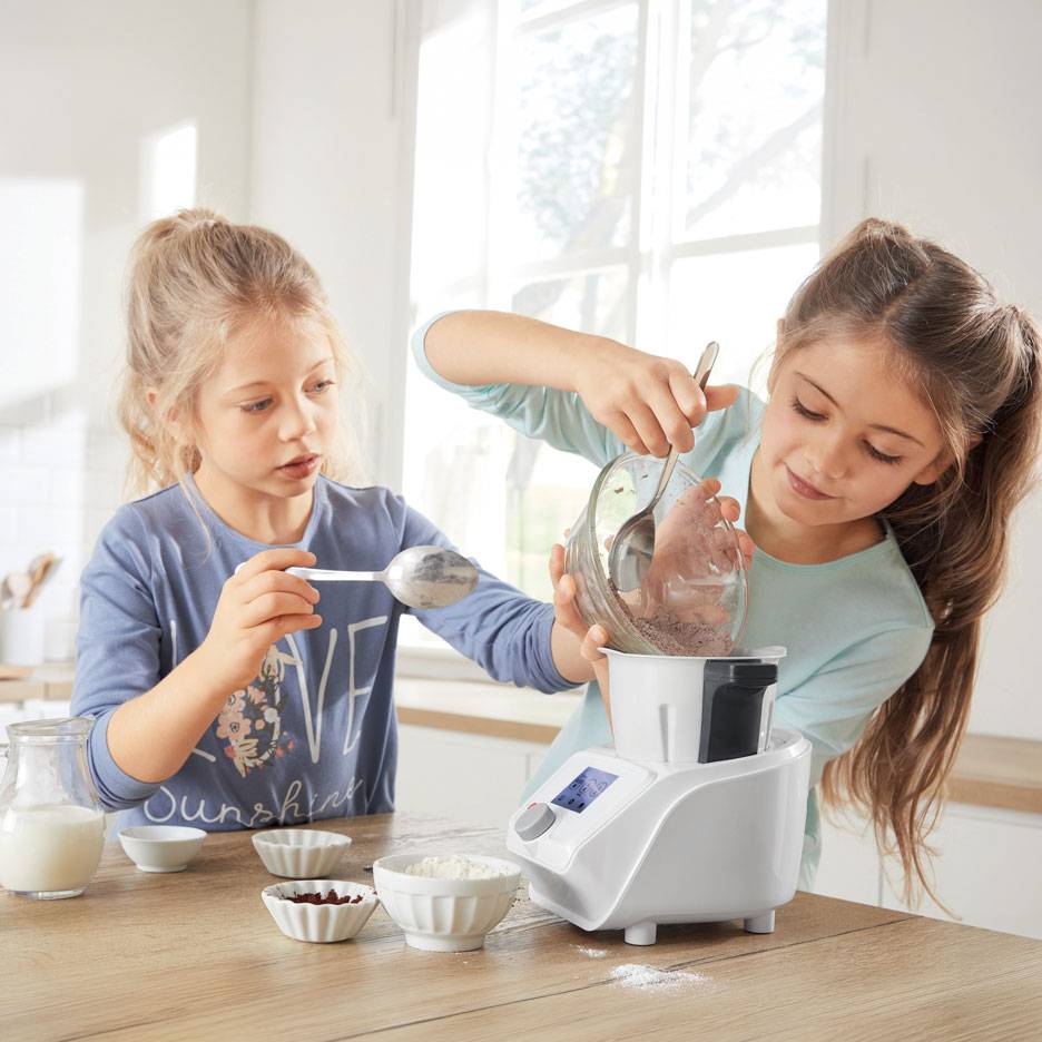 Two girls prepare dough in a kitchen machine, with bowls of ingredients on the table.