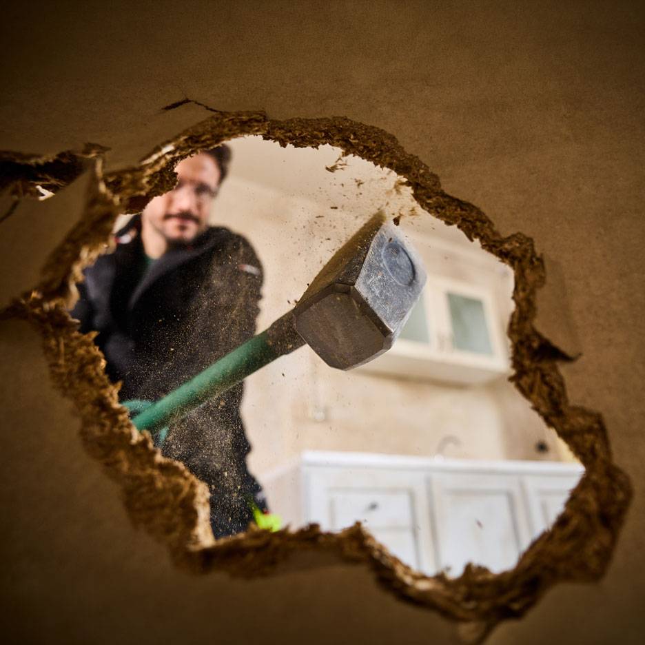 Man with sledgehammer demolishing a wall, seen through a hole, with kitchen cabinets in the background.