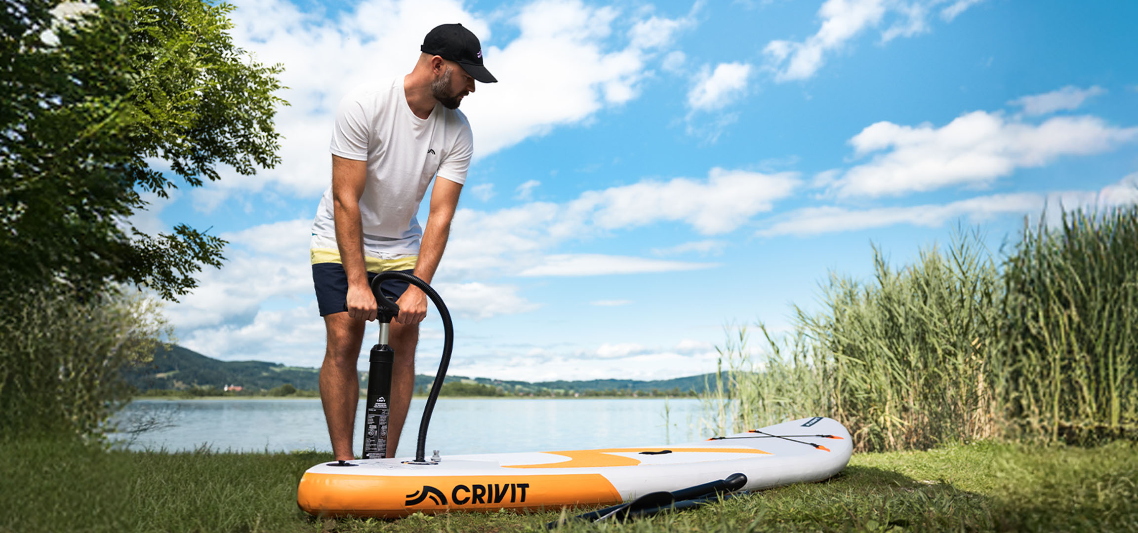 Man inflating CRIVIT SUP board by a lake on green grass.