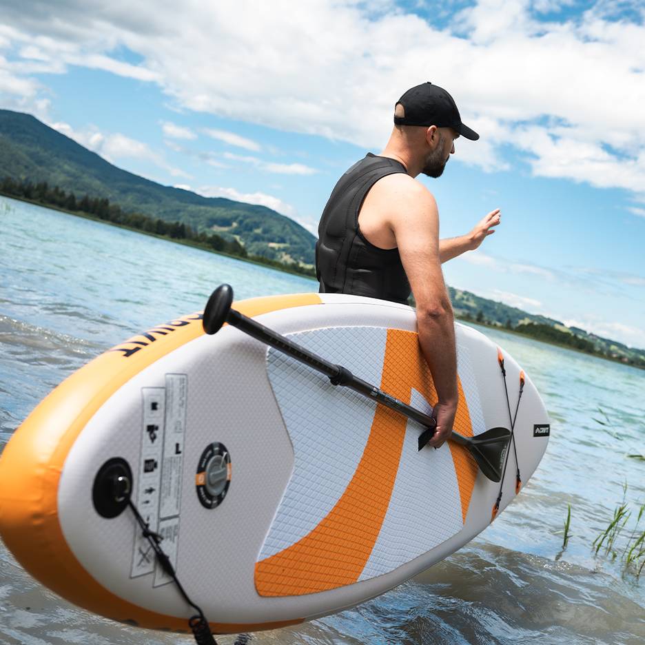 Man in life vest and cap holding an inflatable paddleboard in the lake.