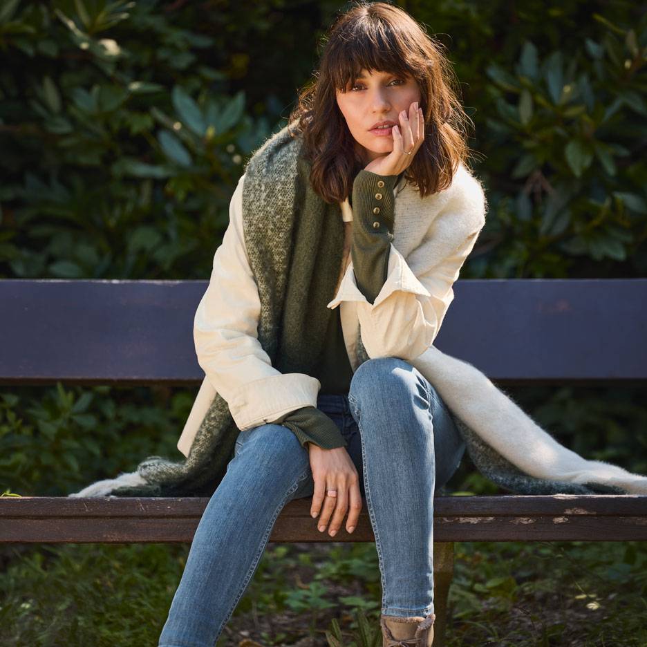 Woman in jeans, white jacket, and green scarf sitting on a park bench.