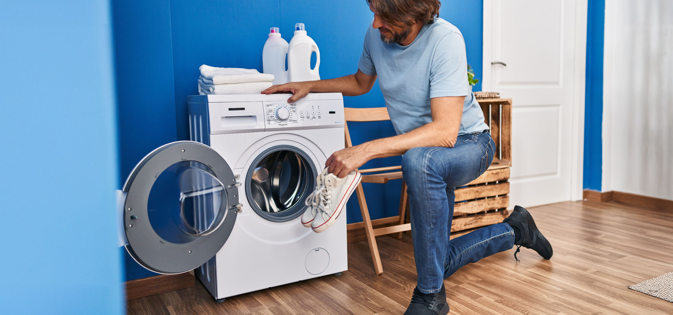 Man putting shoes into washing machine, with detergents and towels nearby.