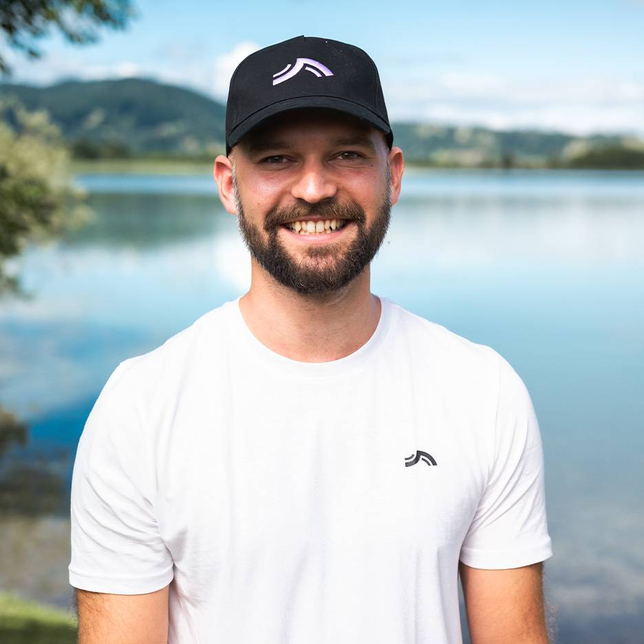 Smiling man in white cap and T-shirt, with a lake and mountains in the background.