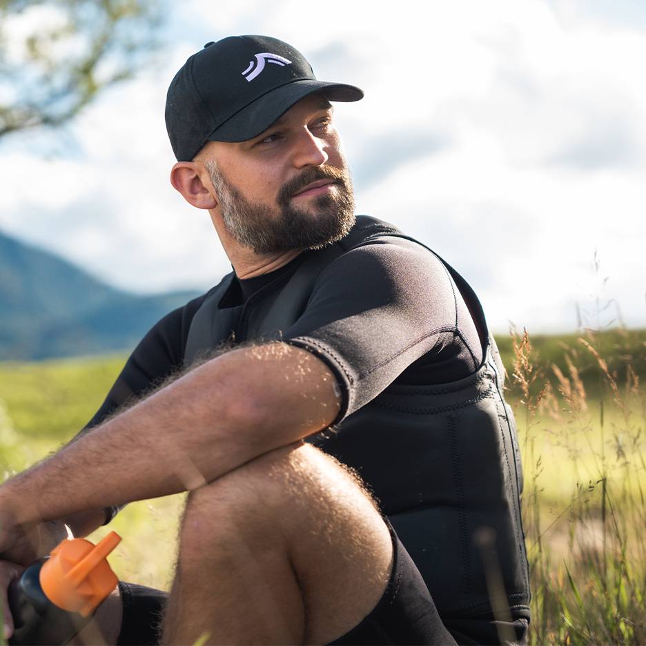 Man in black cap and neoprene vest sits in grass, looking to the side.