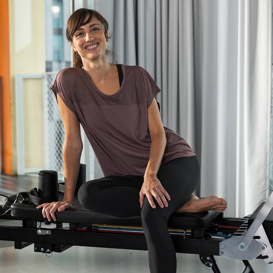 Woman in activewear smiling, sitting on a Pilates reformer machine.