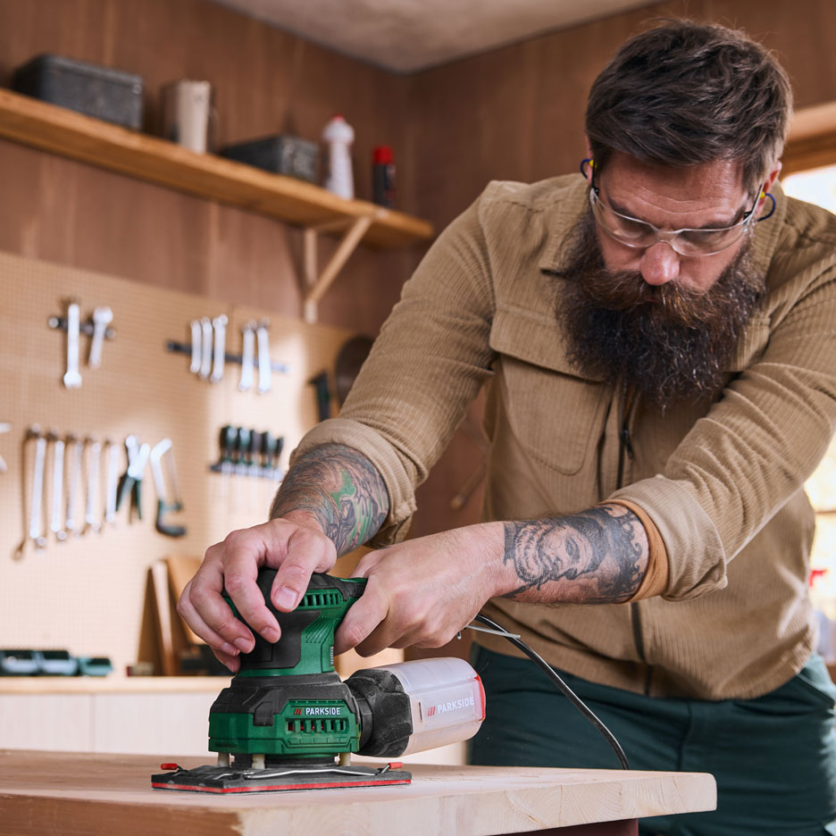Man in workshop sanding a wooden board with an electric sander.