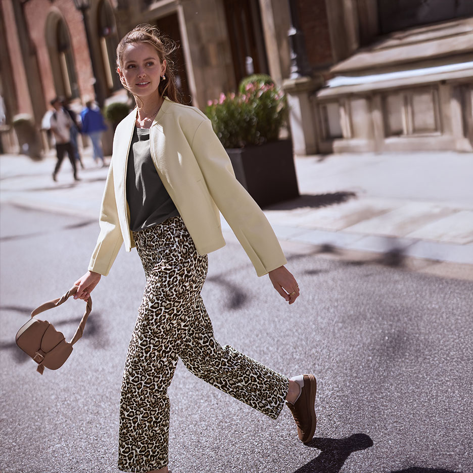 Woman in a beige jacket, green t-shirt, and leopard print trousers, walking down a street with a handbag.