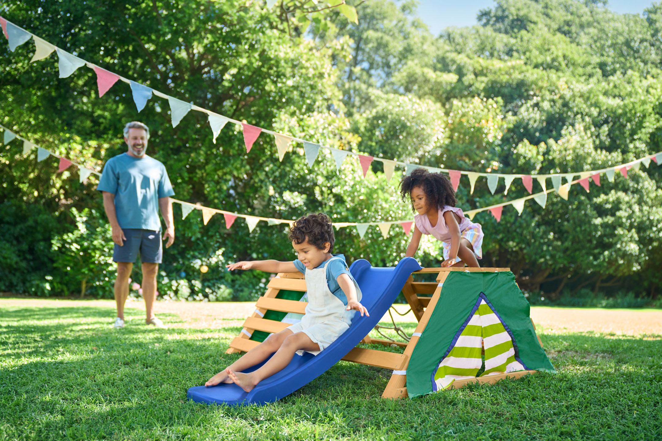 Children playing on a wooden climbing frame with slide and tent in a garden with bunting.