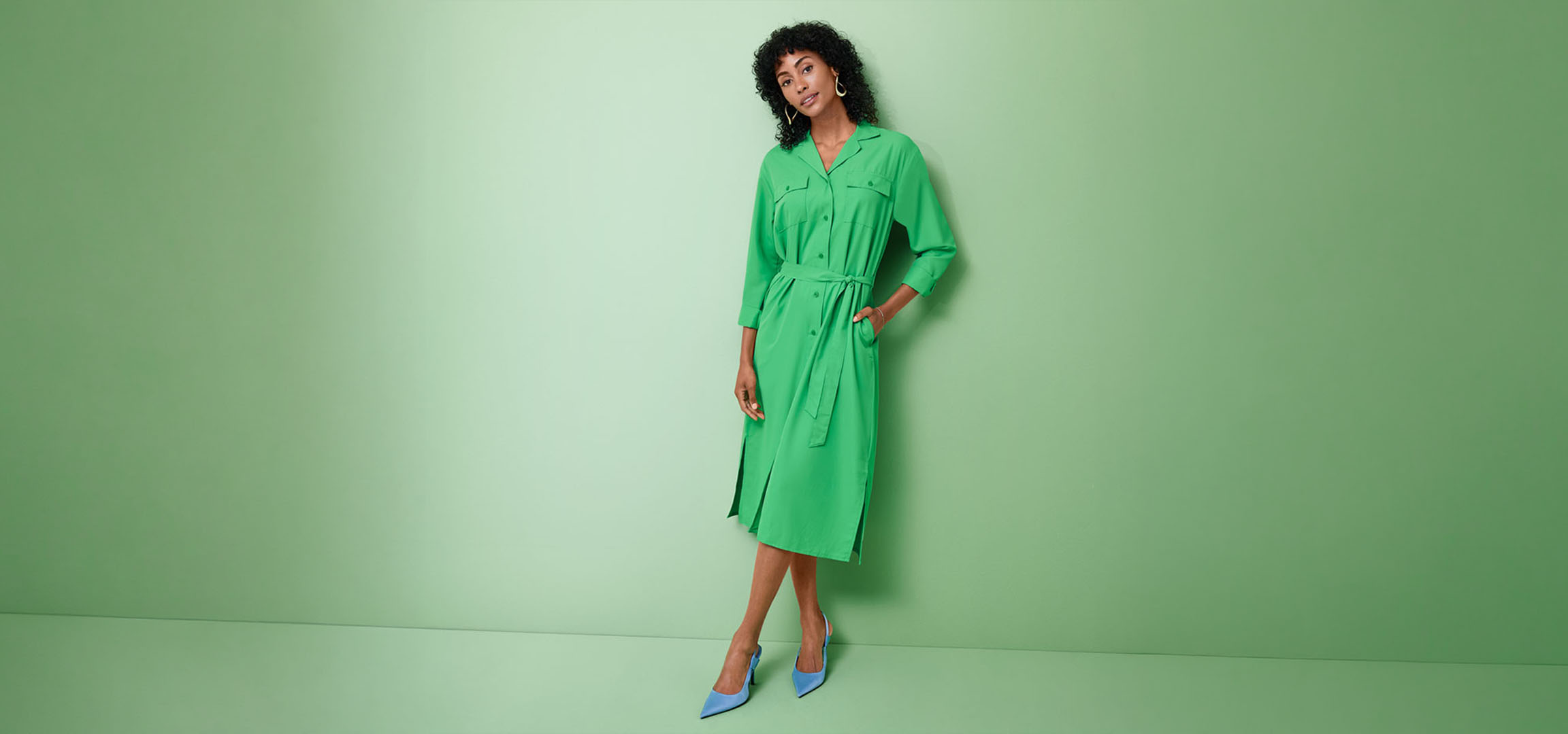 Woman in a green shirt dress and blue heels, posing against a green background.