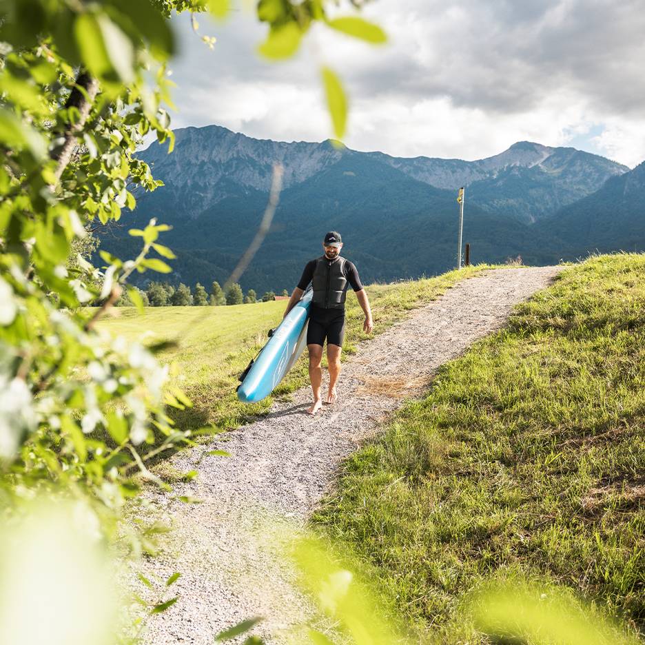 Man in wetsuit and life vest carrying a paddleboard in a mountainous landscape.