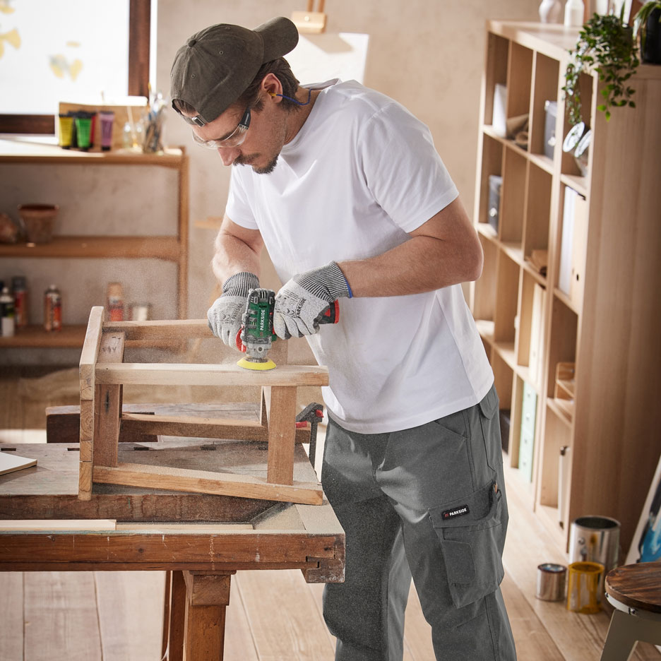 Man sanding wooden furniture with a power tool, wearing work trousers and protective gear.