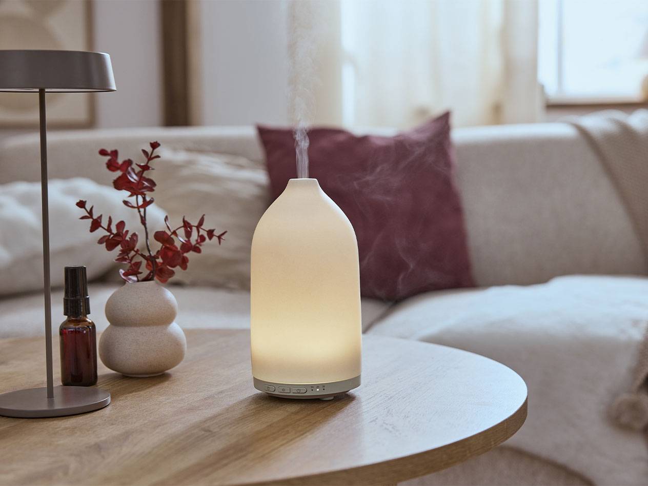 Illuminated aroma diffuser, oil bottle, and vase with branches on a table.