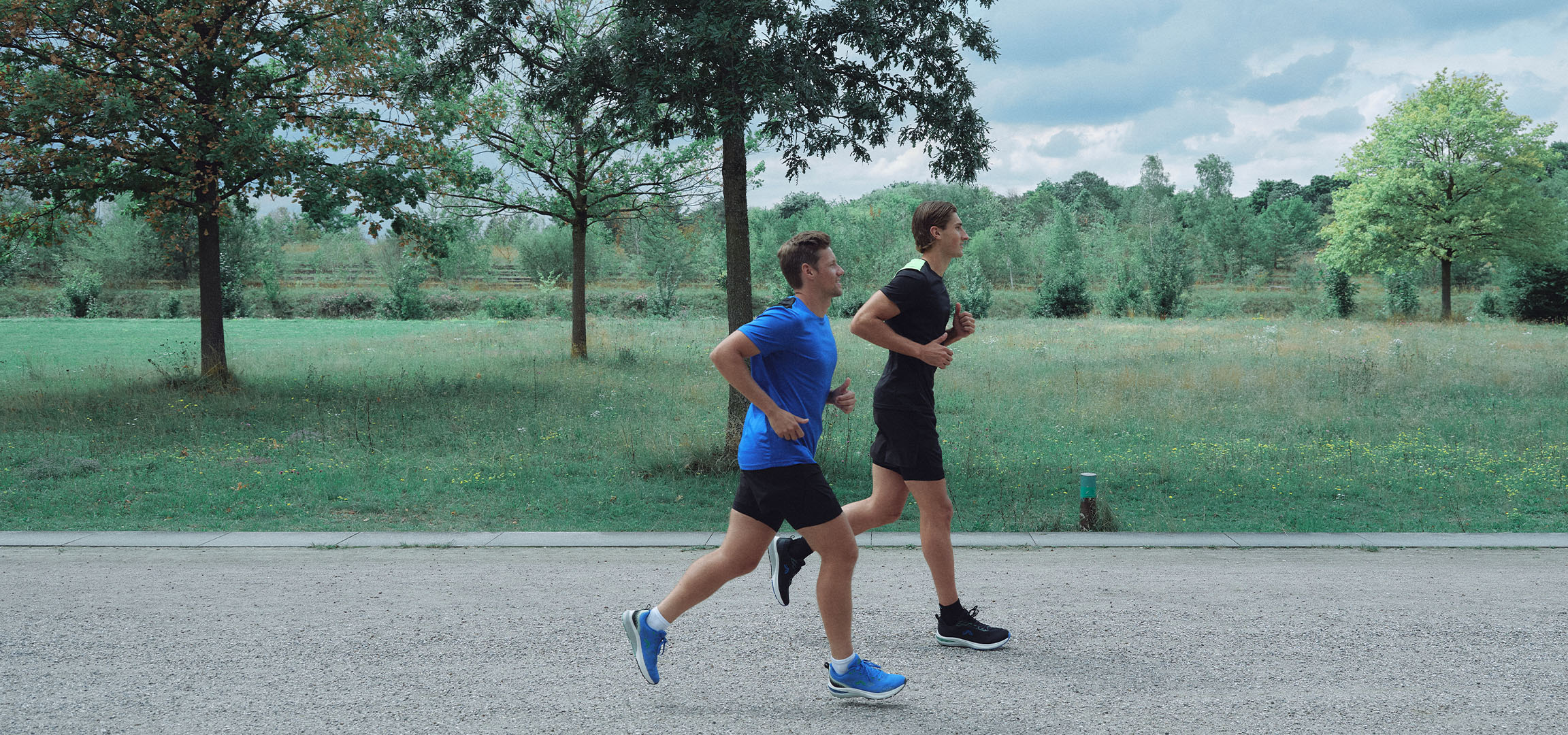 Two men running on a gravel path in a park, dressed in sportswear.