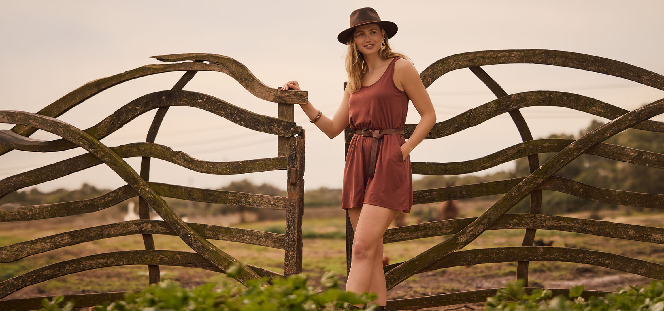 Woman in dress and hat, leaning on a wooden fence in the countryside.