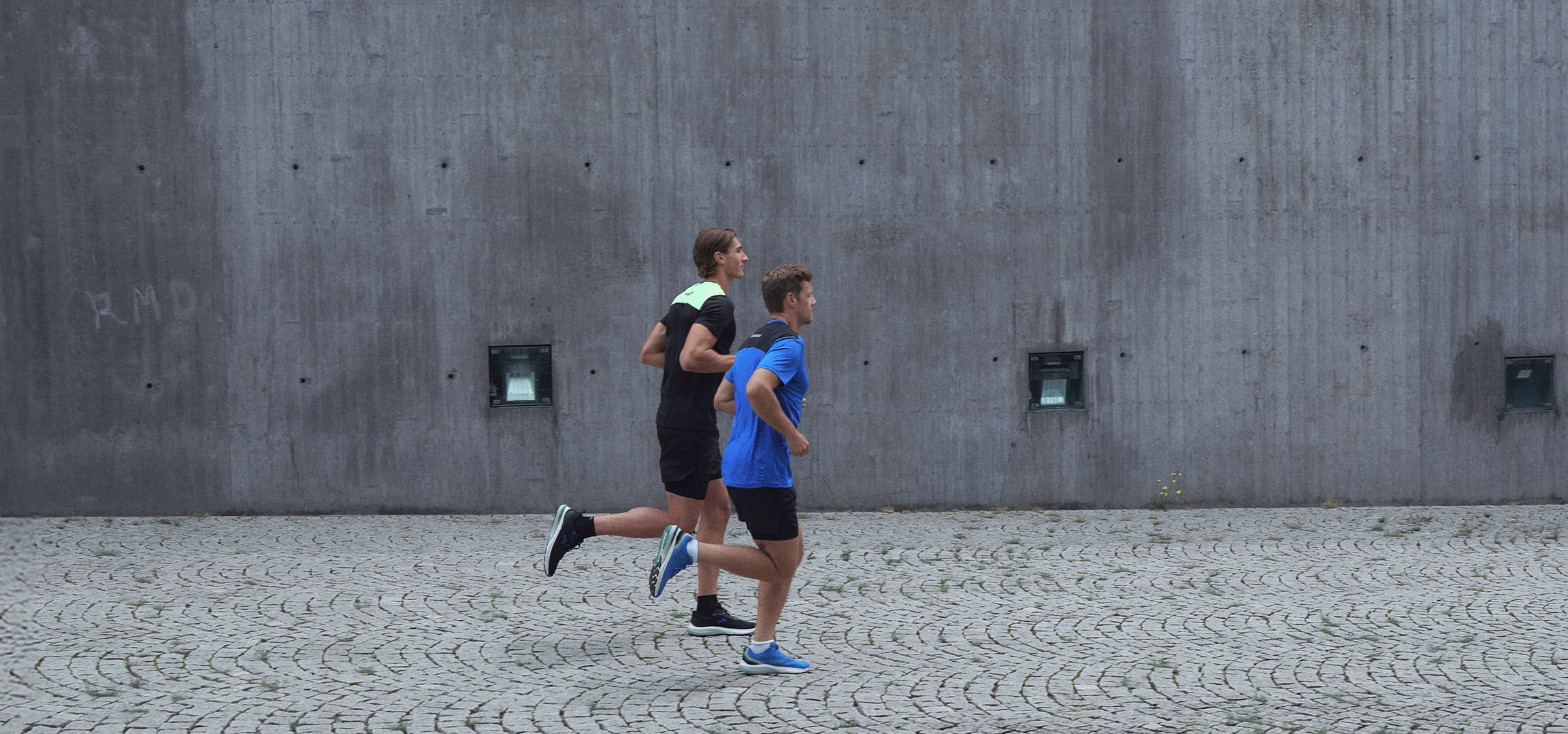 Two men in sportswear running along a wall.