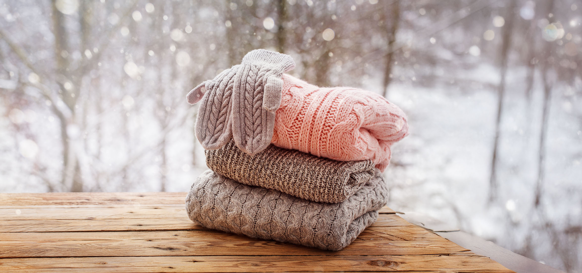 Stack of knitted sweaters and mittens on a wooden table with a snowy background.