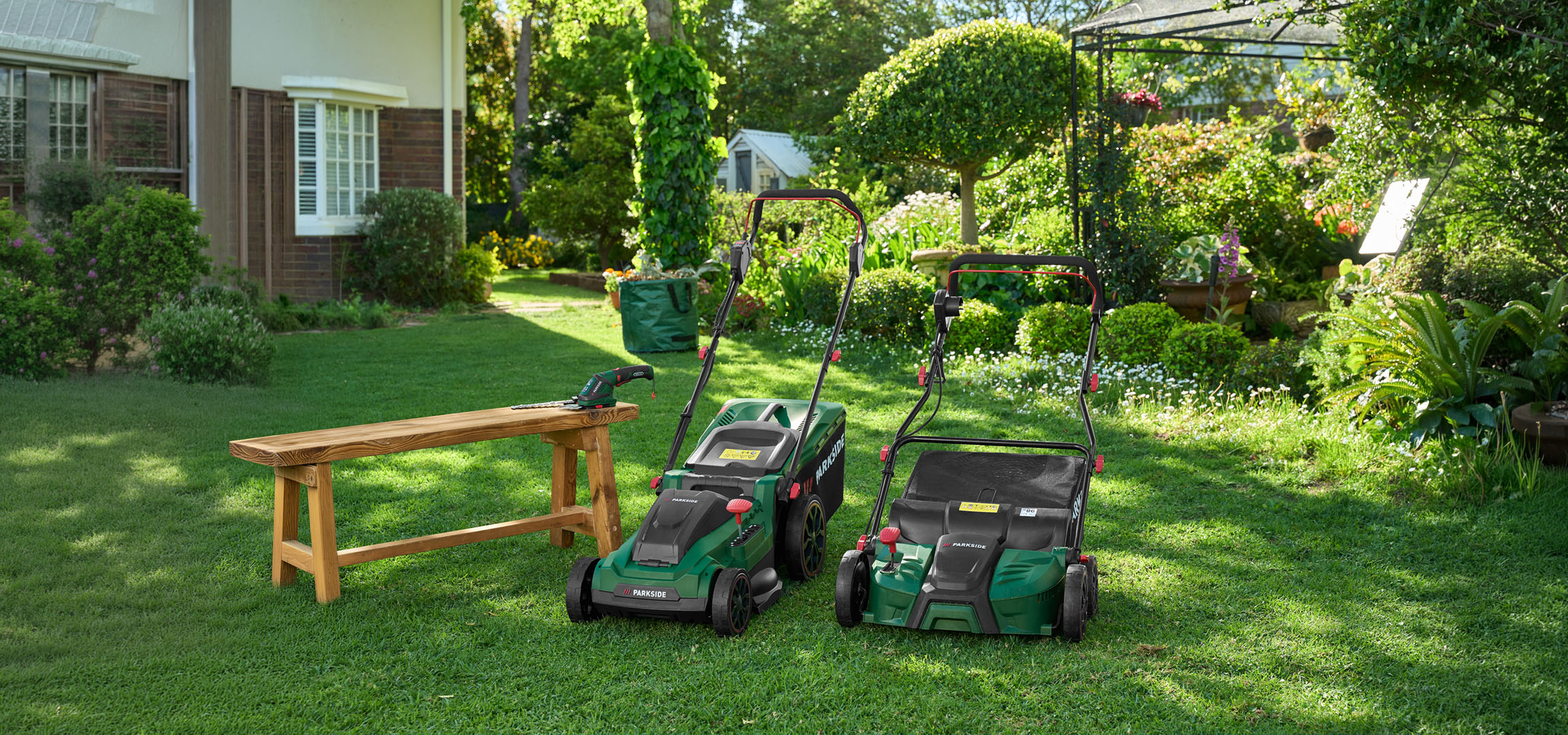 Two lawnmowers and a hedge trimmer in a lush green garden.