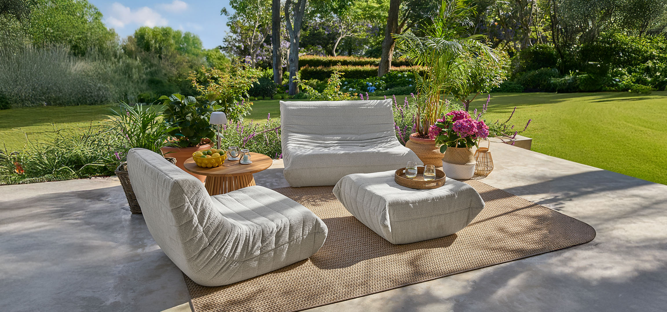 Light grey armchairs and poufs on a terrace with a wooden table and green lawn.