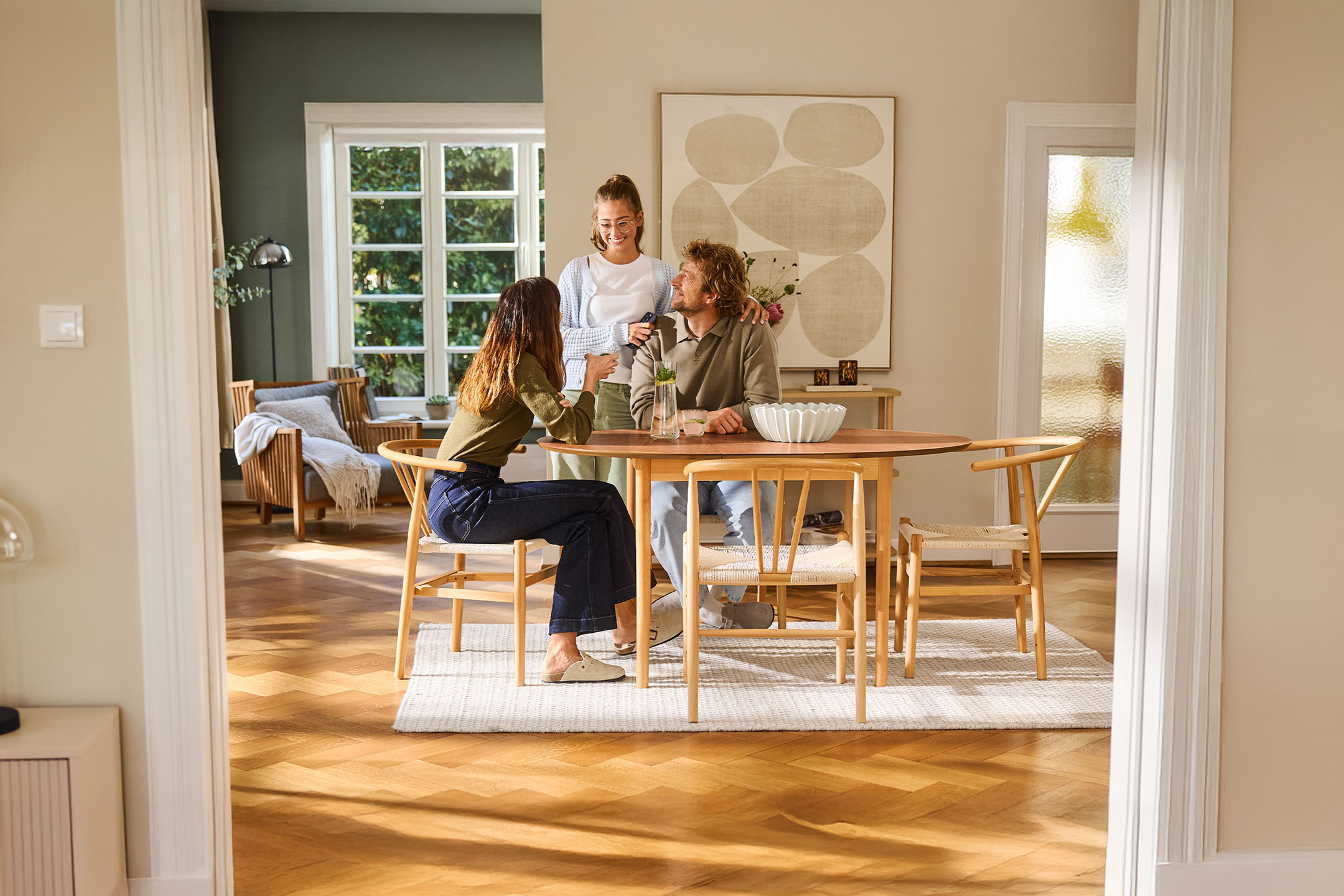 Three people enjoy a meal at a wooden table in a bright dining room.