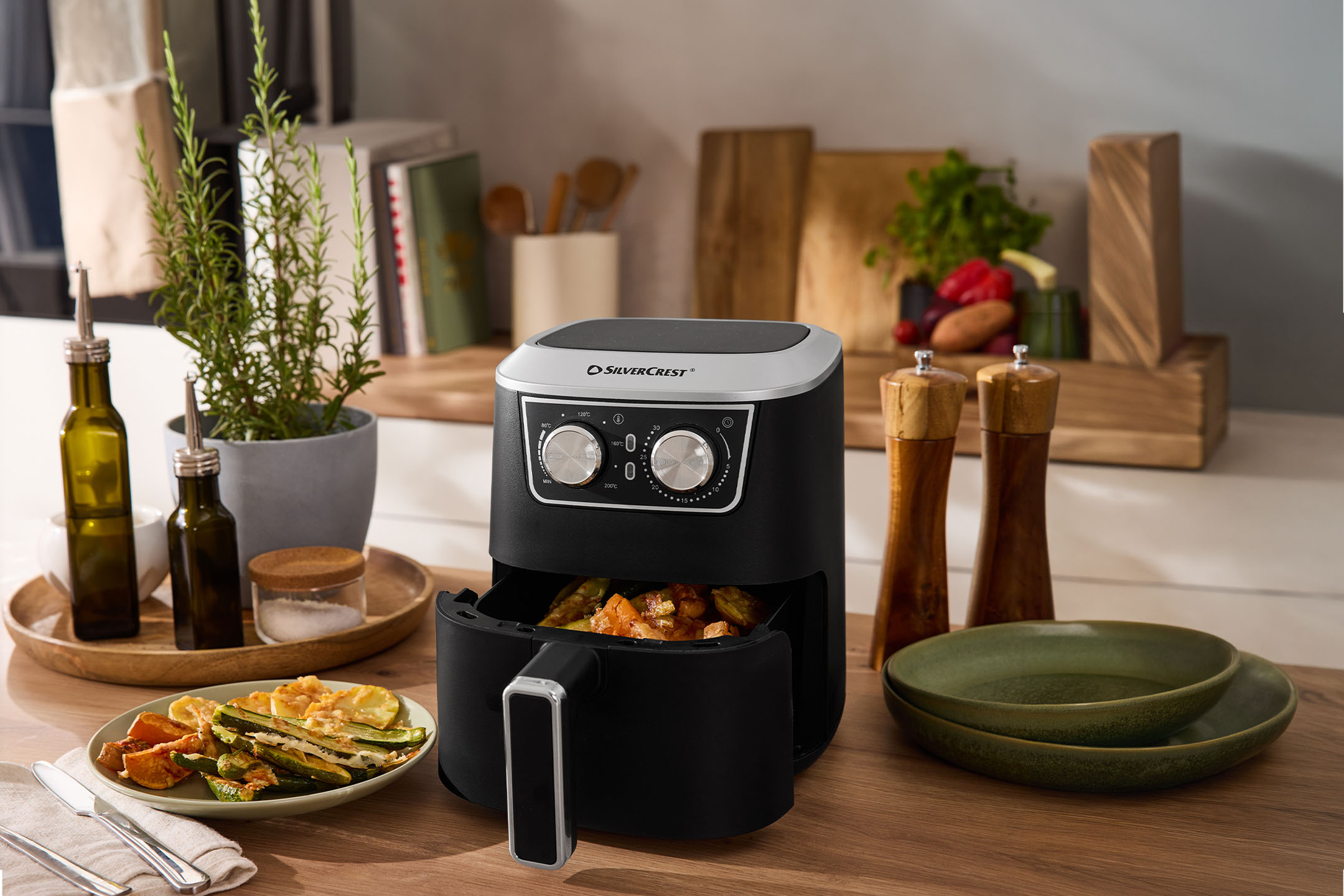 Air fryer with roasted vegetables, olive oil, salt, and pepper on a kitchen counter.