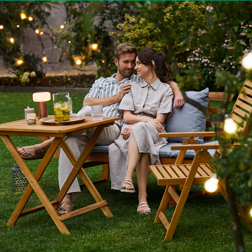 Couple relaxing on wooden garden furniture with drinks and candles.