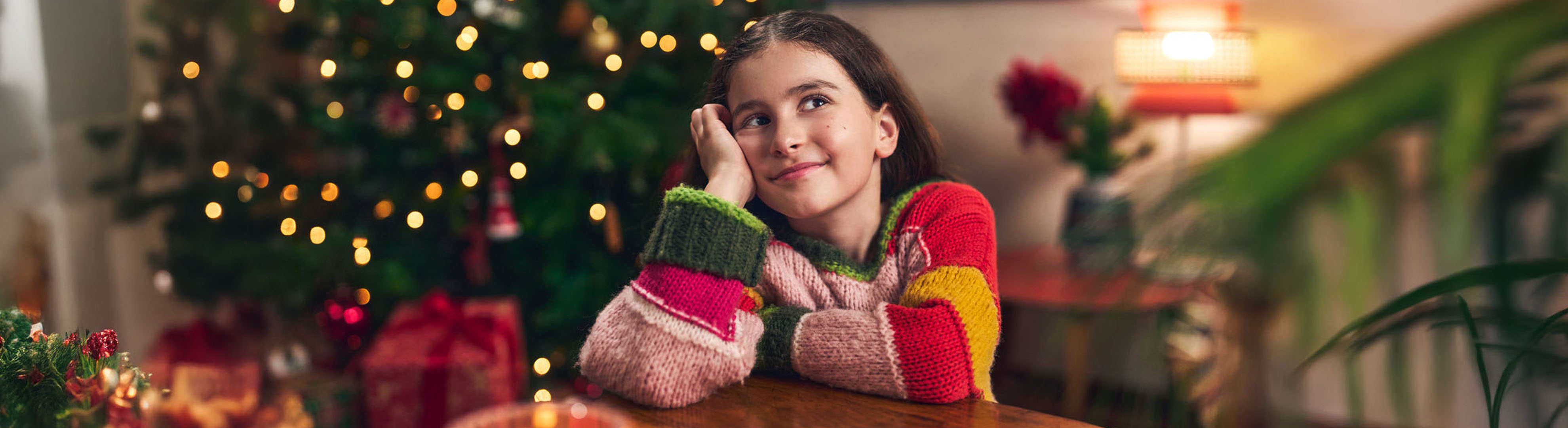 Smiling girl in colorful sweater, leaning on a table with a Christmas tree in the background.