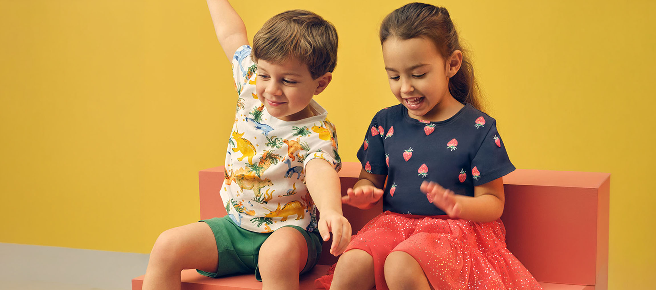 Two children, a boy and a girl, sitting on a pink bench wearing summer clothes.