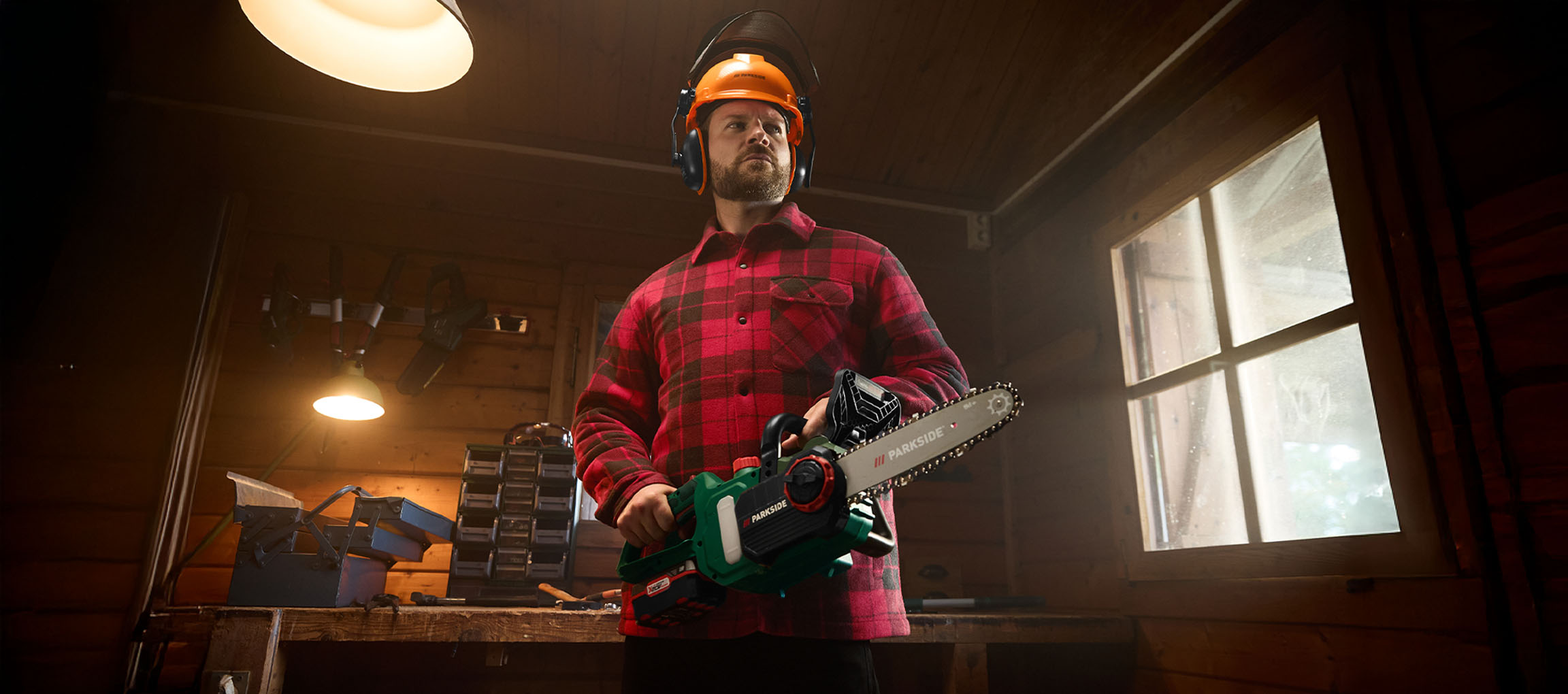 Man in plaid shirt and safety helmet, holding a Parkside chainsaw in a workshop.