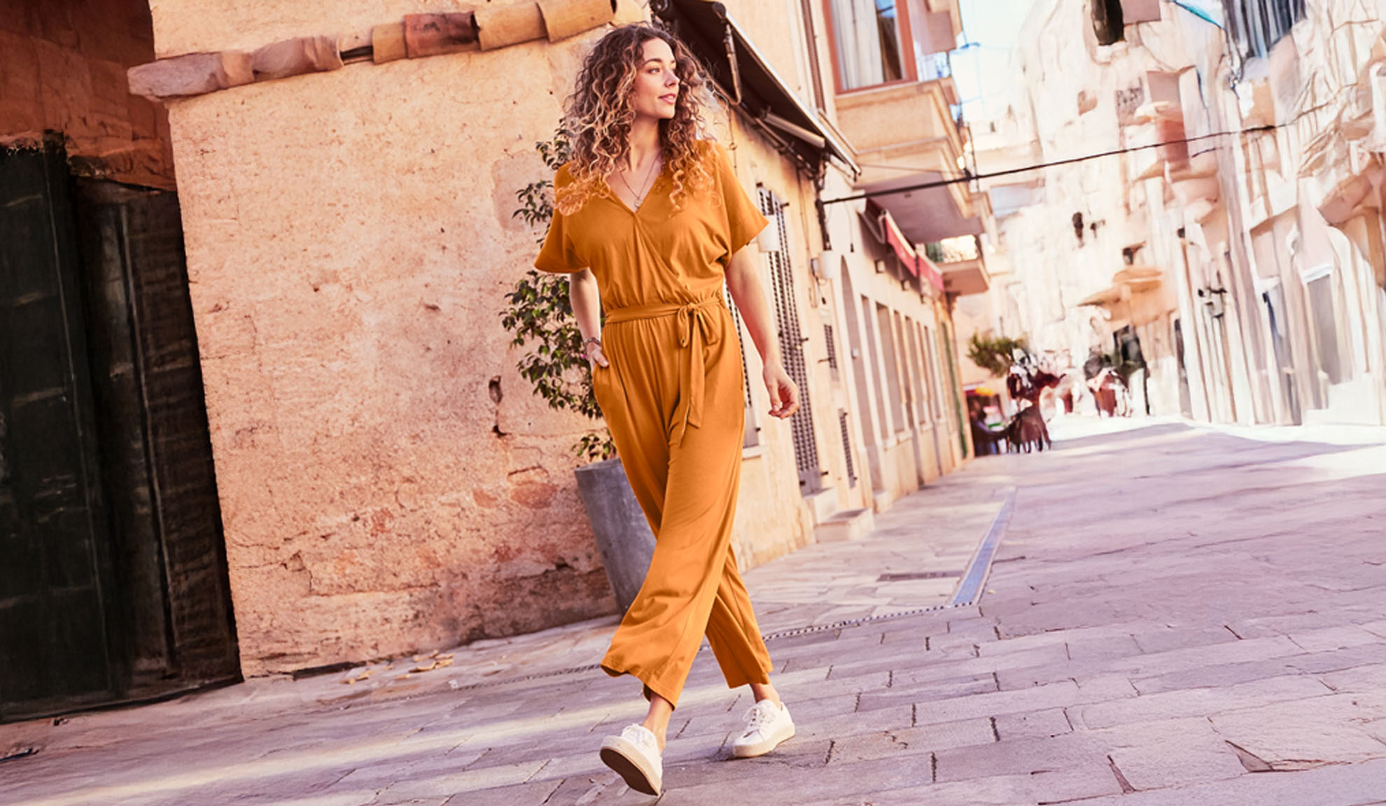 A woman strolls down a sun-drenched cobblestone street wearing a stylish mustard-yellow jumpsuit and white sneakers.