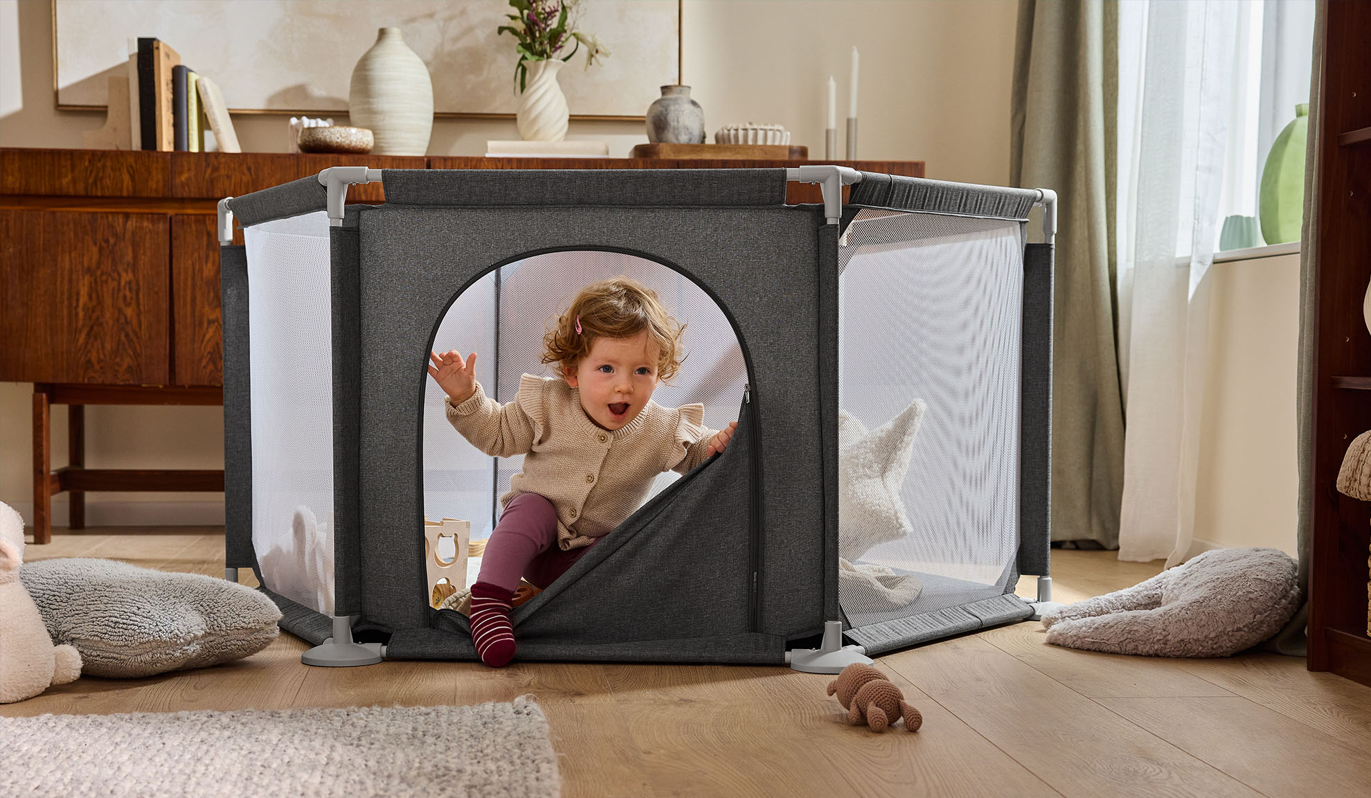 Happy baby crawling out of a grey playpen in a cozy living room.