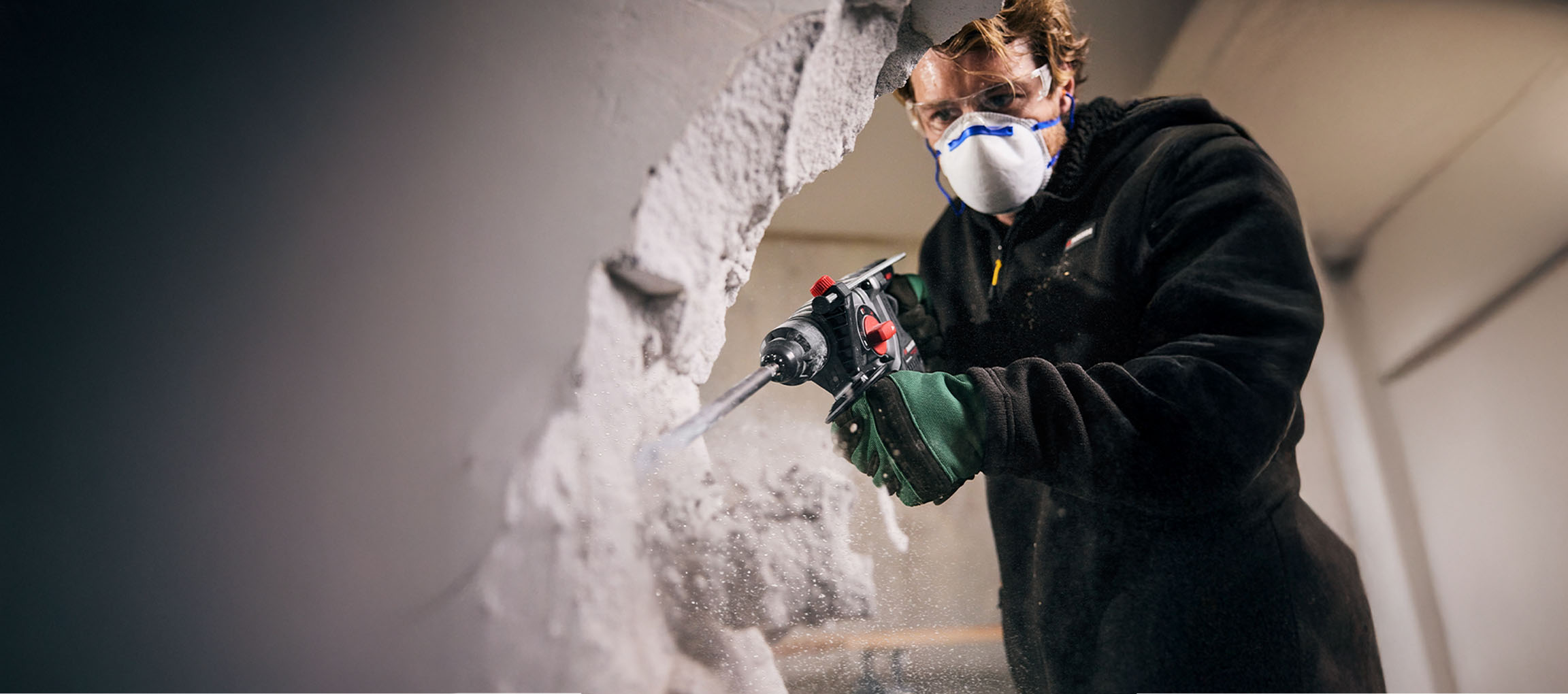 Man in safety glasses and mask using a hammer drill to break through a wall.