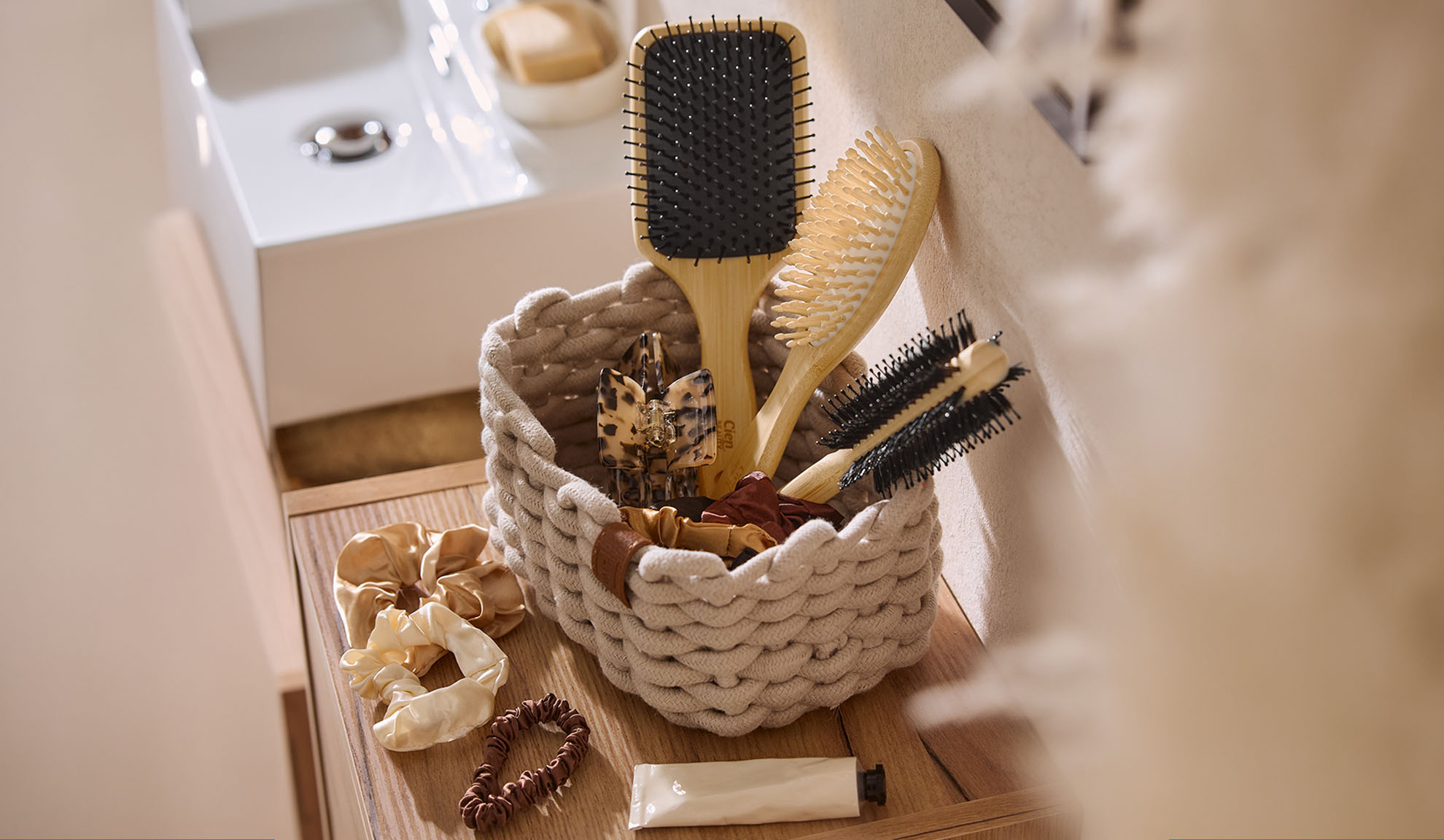 Rope basket with wooden hairbrushes, hair clips, and scrunchies in a bathroom.
