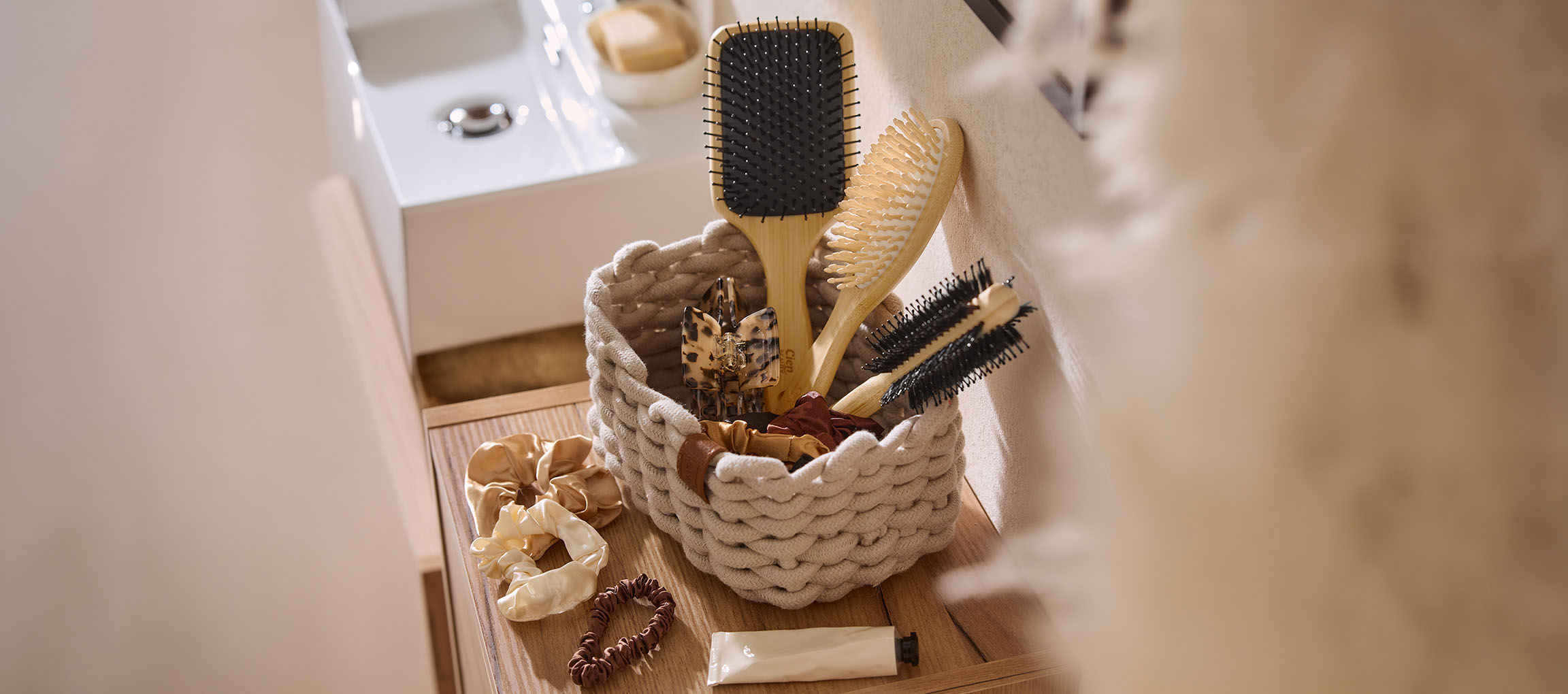 Basket with hair accessories: brushes, clips, and scrunchies, on a wooden bathroom shelf.