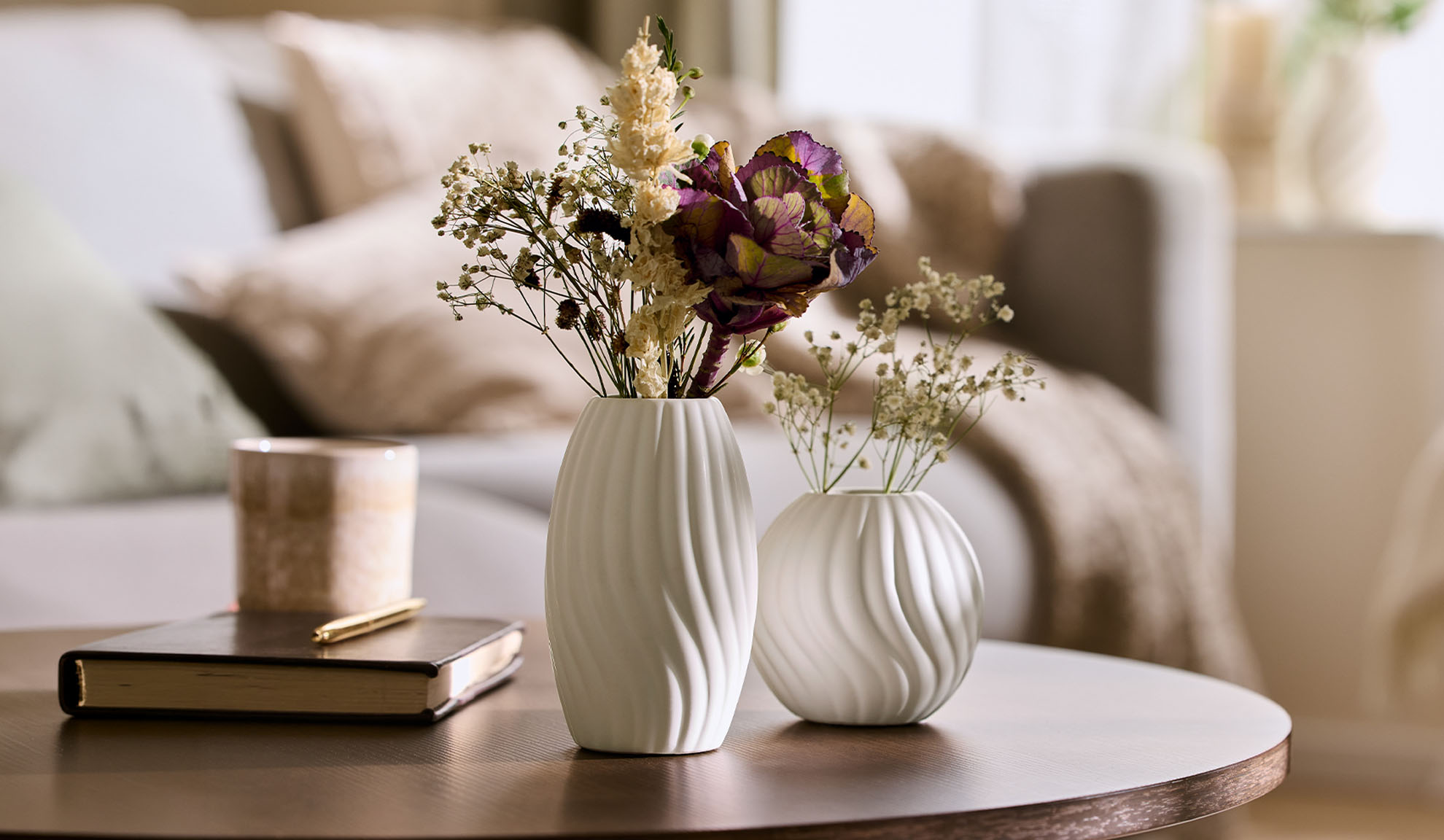 White vases with dried flowers on a wooden coffee table in a cozy living room.