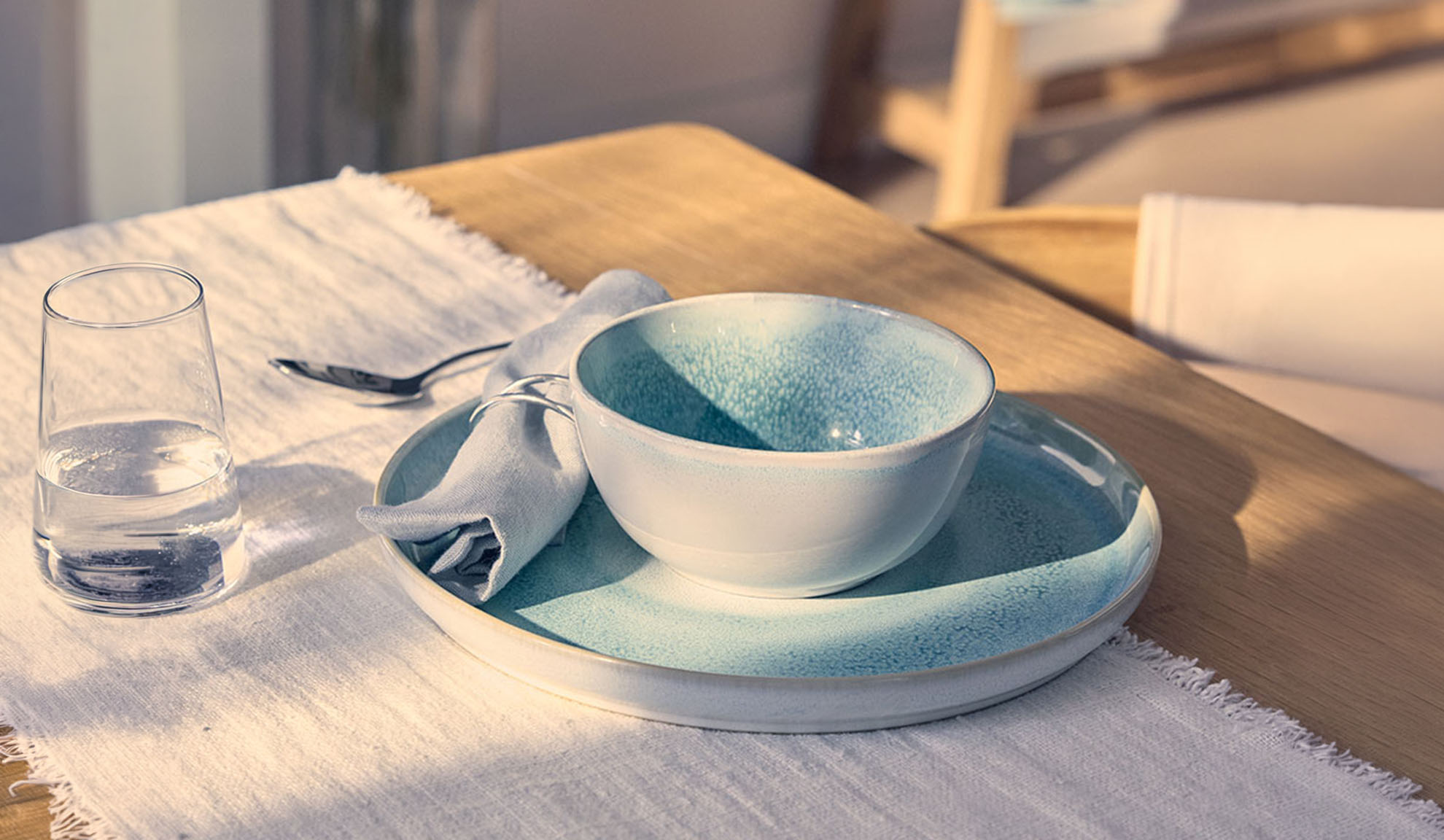 Table setting with a blue bowl, plate, glass of water, and napkin on a wooden table.