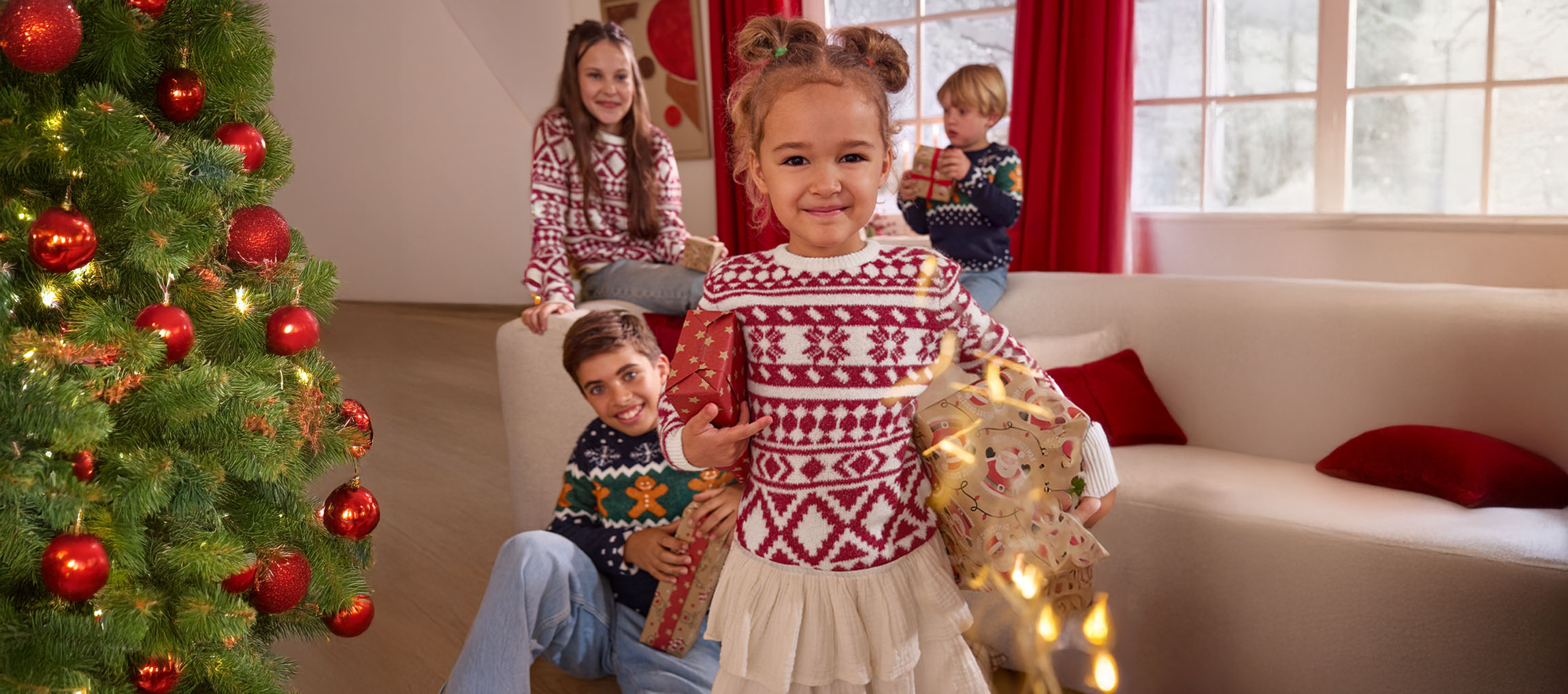 Children in festive Christmas sweaters holding gifts next to a decorated Christmas tree.