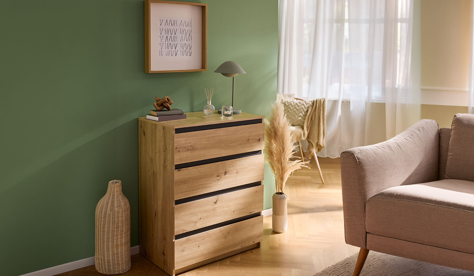 Wooden chest of drawers, table lamp, and beige sofa in a living room with a green wall.